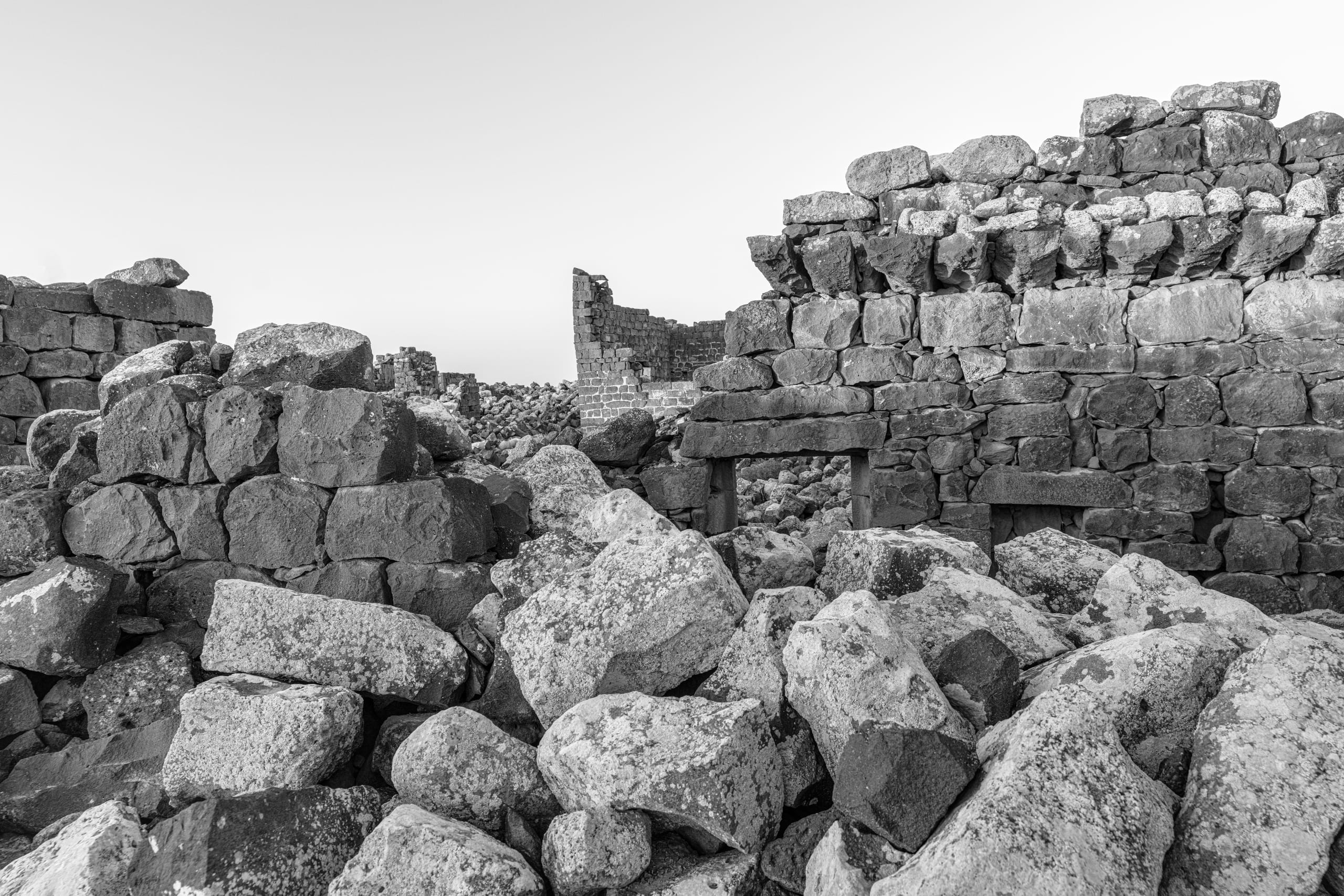 Monochrome photograph of crumbling stone ruins and jumbled basalt boulders, highlighting rough textures, broken walls, and weathered masonry against a pale sky.