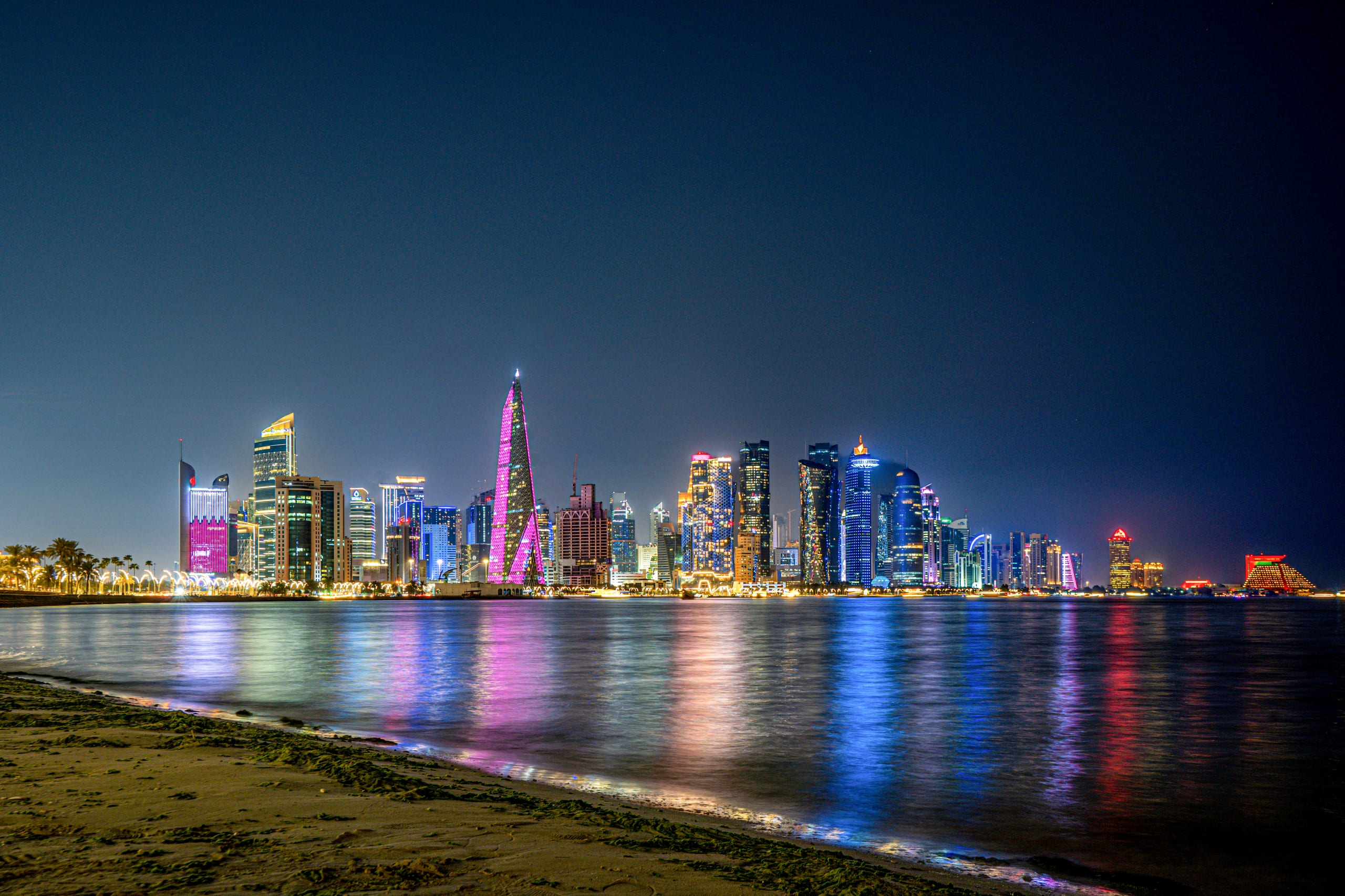 A brightly lit skyline of modern skyscrapers at night viewed across a body of water, with colorful light reflections shimmering on the water and a sandy foreground beach.