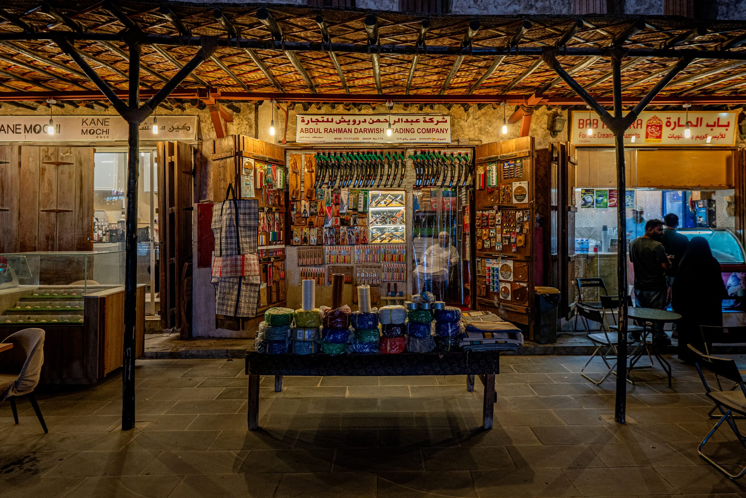 A wide nighttime shot of a traditional storefront with a merchant seated inside among hanging tools. In the foreground, a wooden table holds stacks of colorful wrapped items, beneath a timber-framed woven roof overhang.