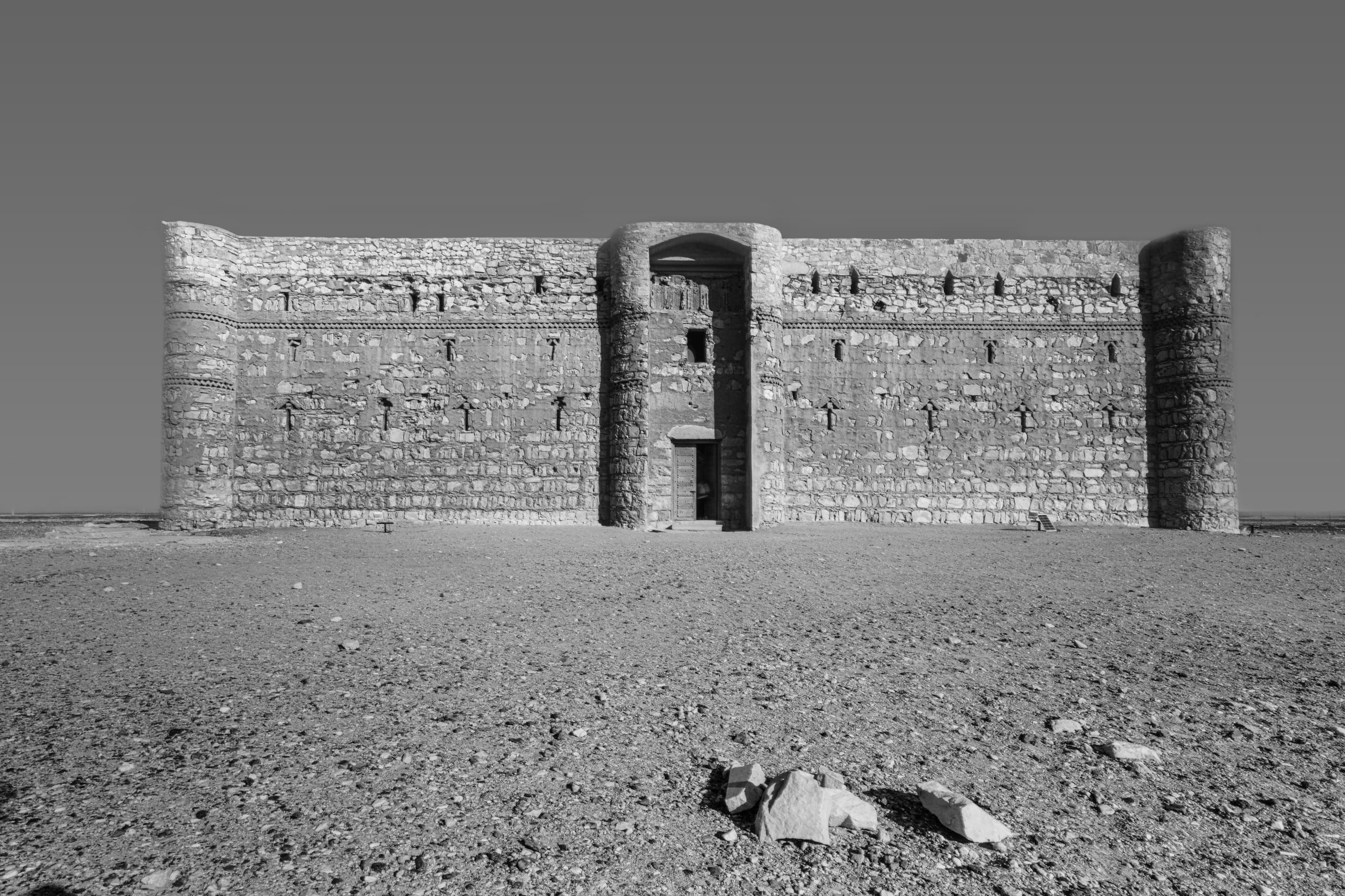 Symmetrical black and white photograph of an ancient stone fortress facade with corner towers and a central arched entrance, viewed straight-on across a flat, gravel-strewn desert floor.