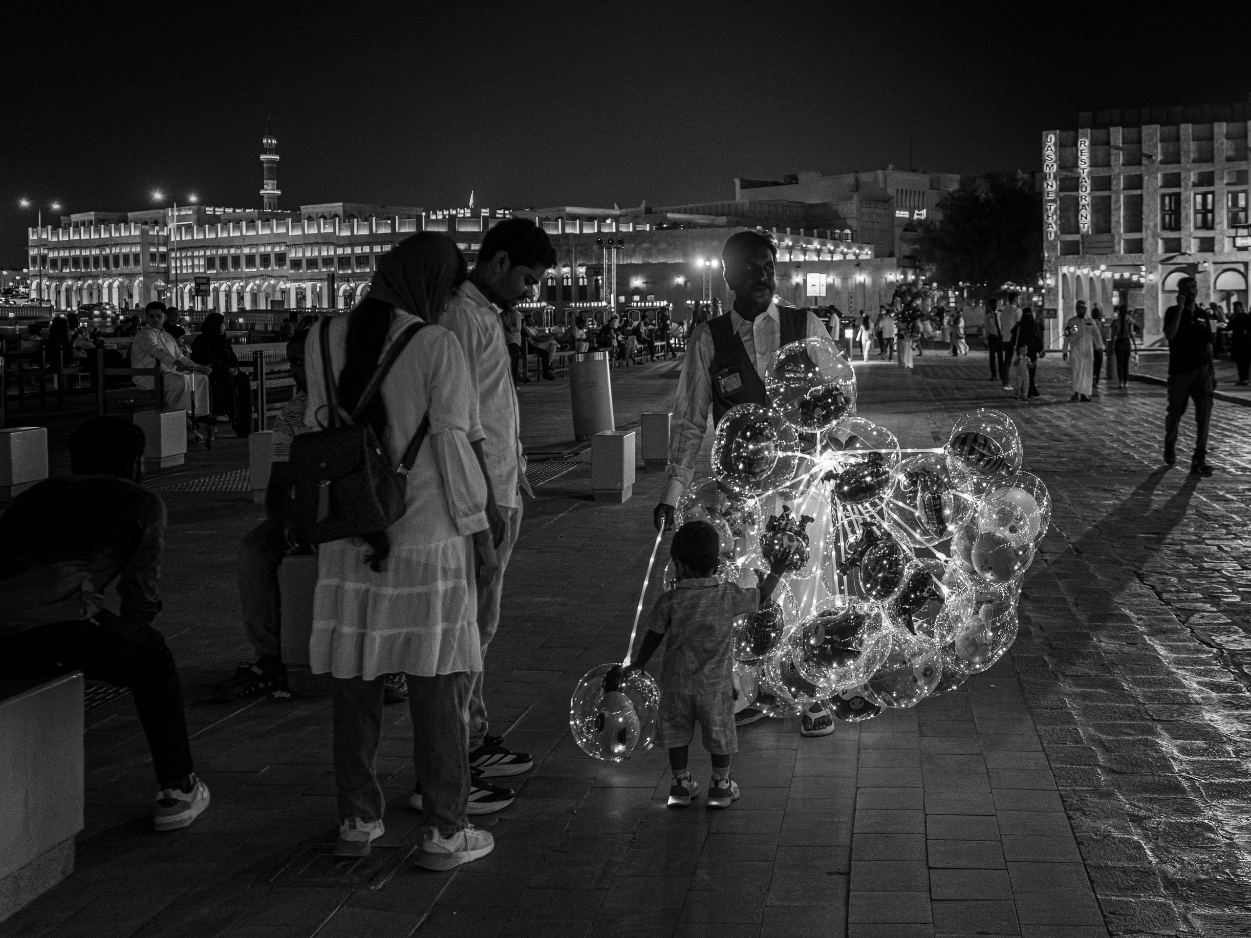 Monochrome street scene at night showing a vendor holding a cluster of glowing spherical balloons on sticks, with a small child reaching up to touch them and several pedestrians watching nearby on a paved plaza.