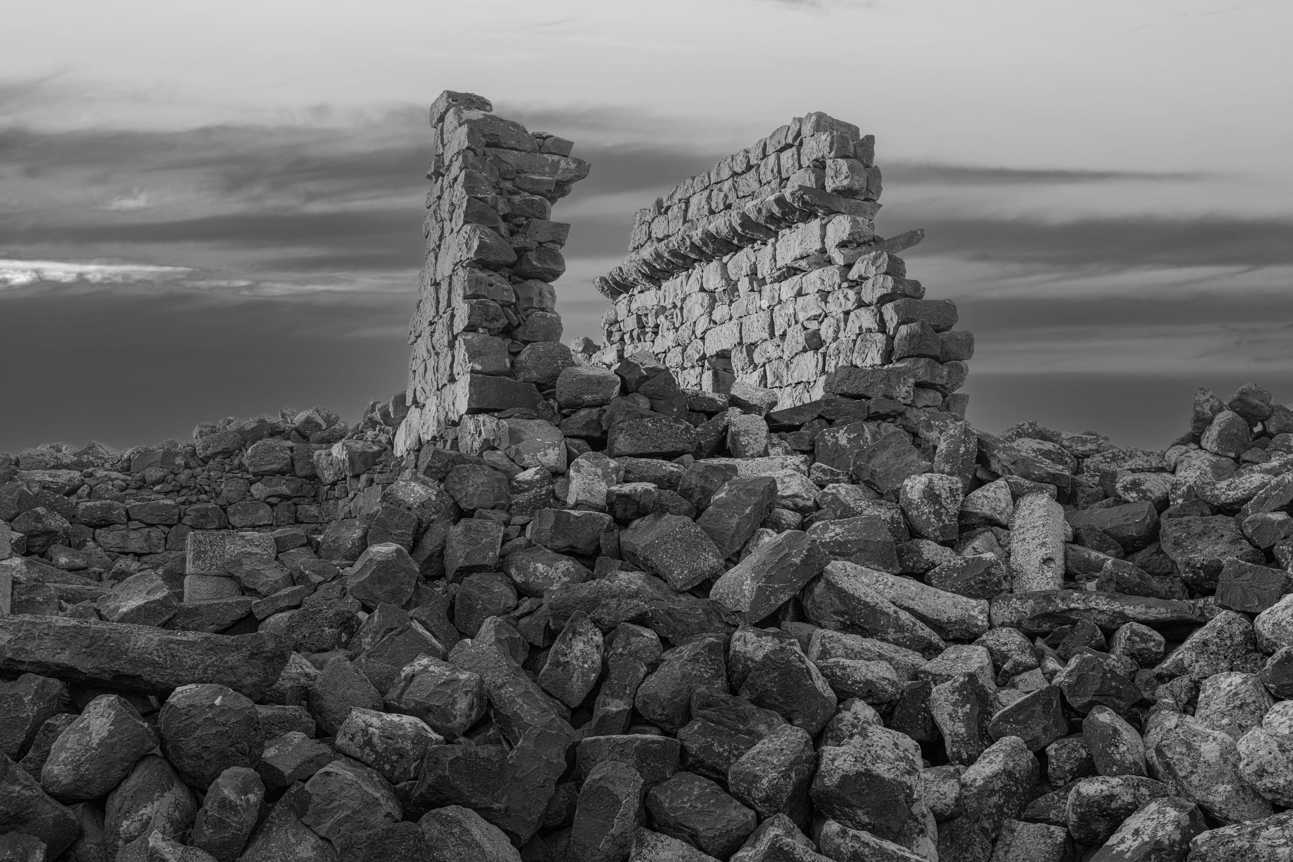 Black and white photograph of ancient stone ruins and collapsed basalt walls surrounded by rubble, under a dramatic cloudy sky.