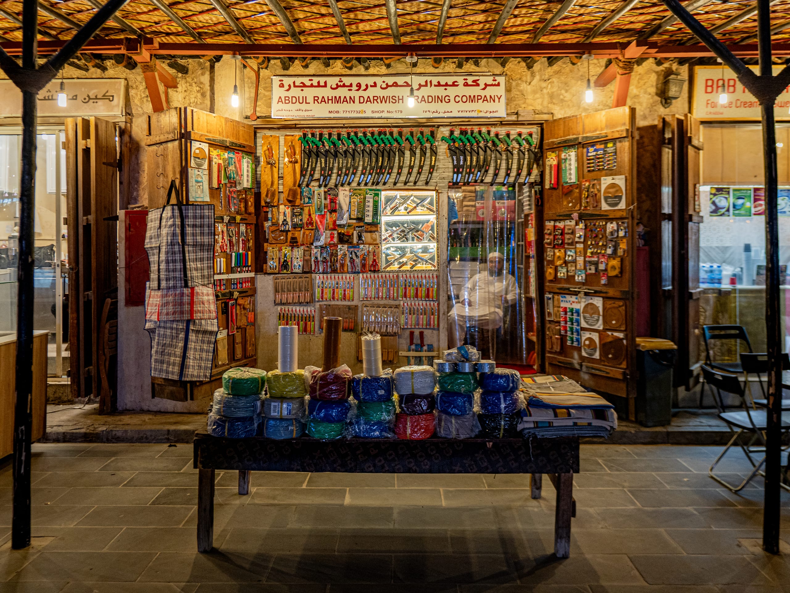 A store facade displaying rows of saws and tools with a seated figure inside, and a wooden table piled with wrapped bundles of goods in the foreground.