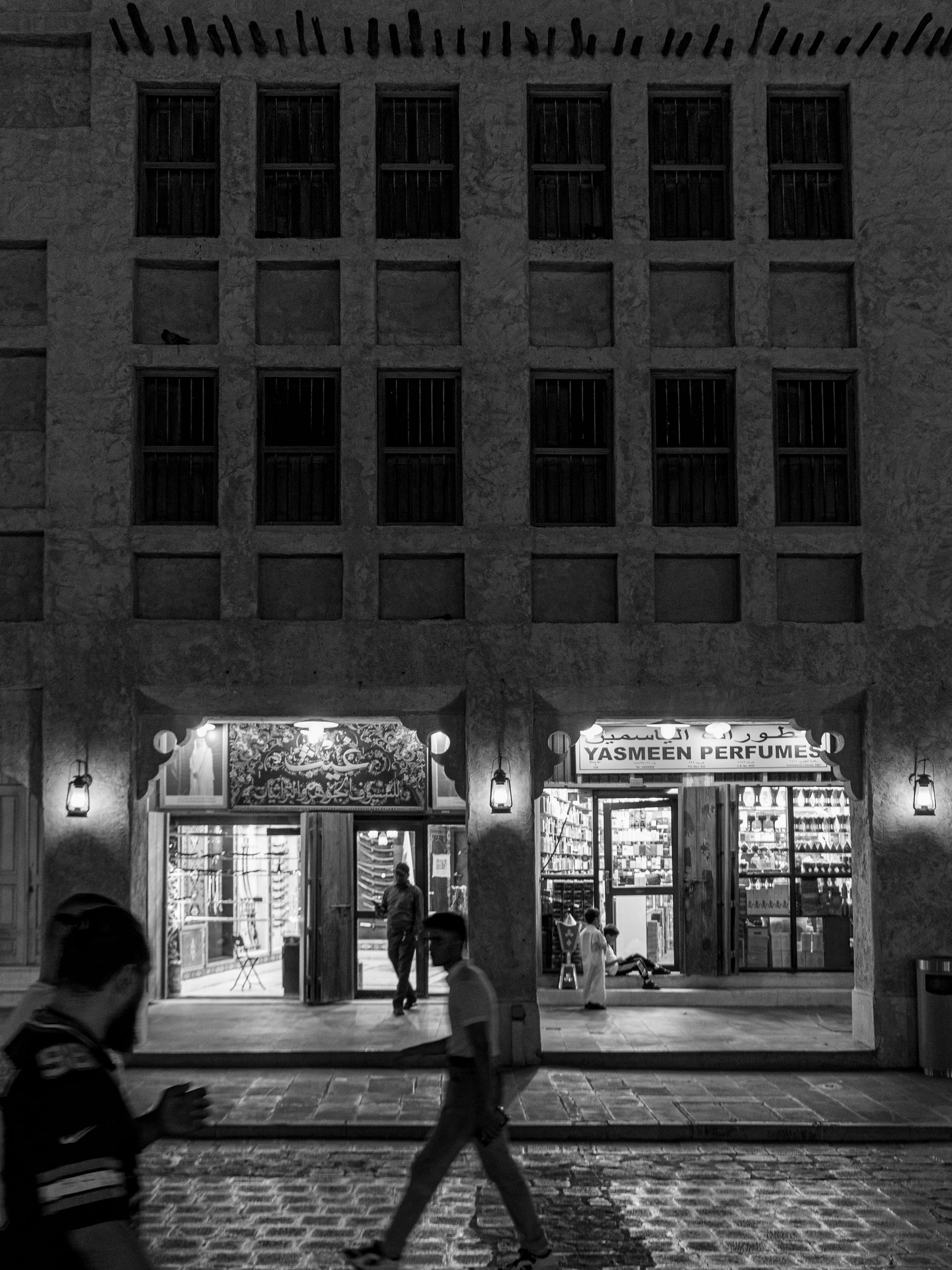Monochrome street-level view of a traditional stone building facade at night featuring glowing shopfronts, hanging lanterns, and blurred pedestrians in motion across a paved walkway.