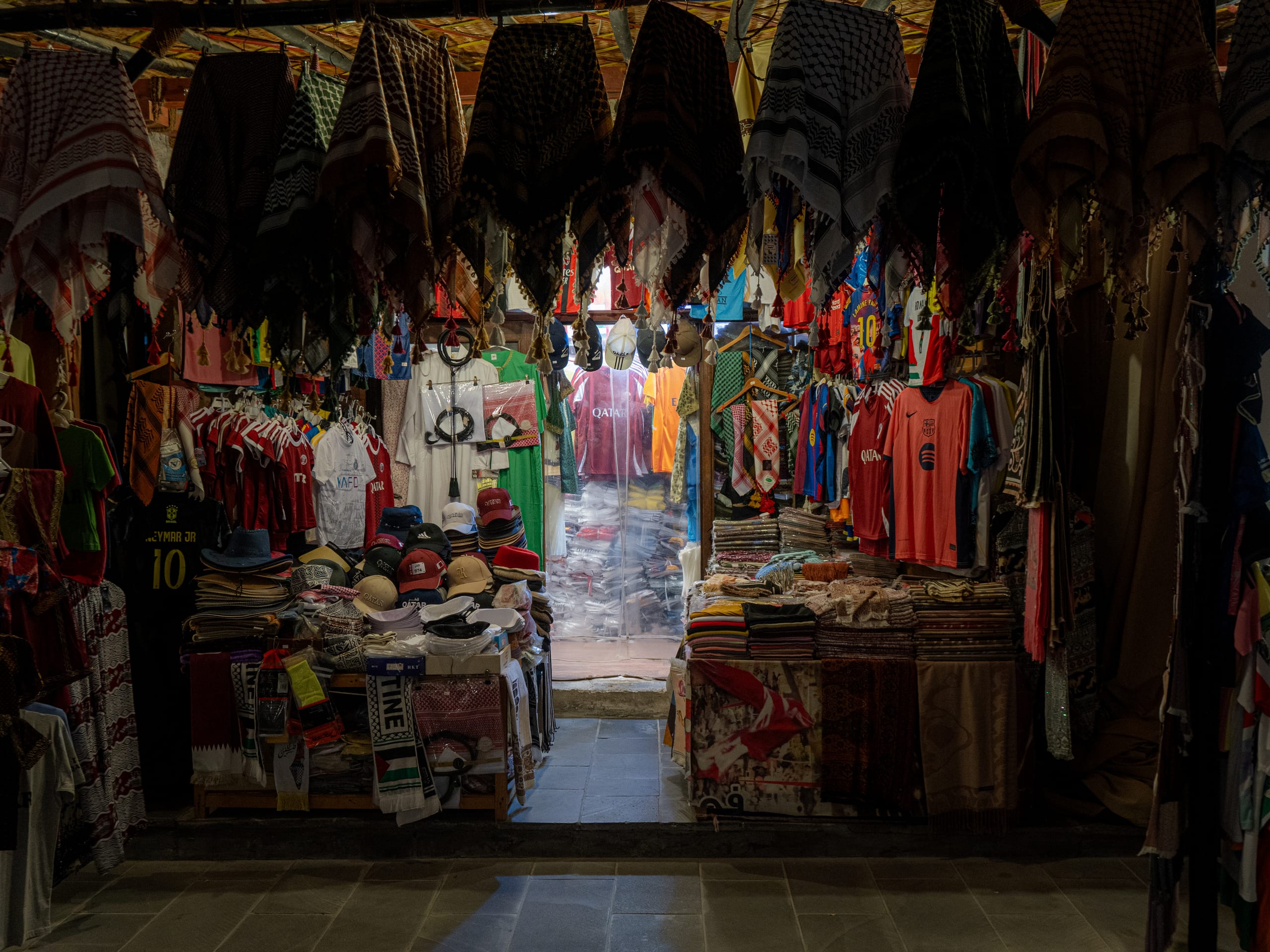 A market stall laden with hanging patterned headscarves, folded textiles, and sports jerseys, viewed from a stone-paved floor looking inward toward a bright open doorway.