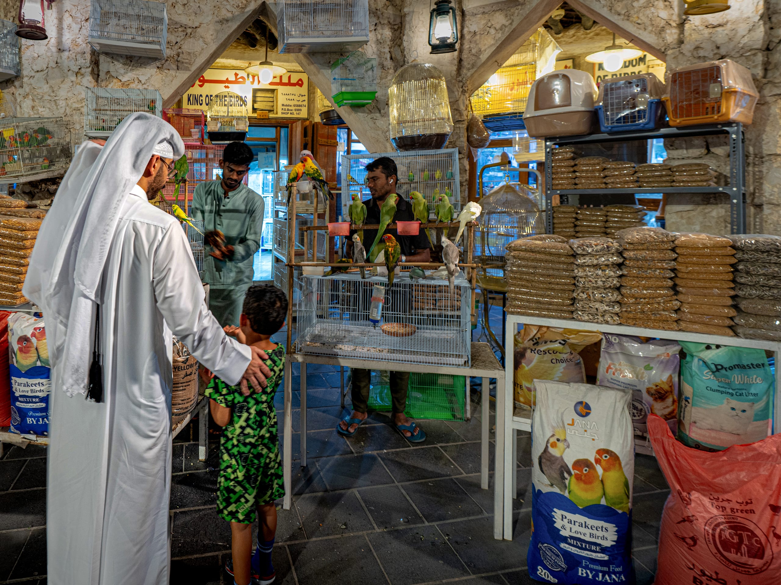 An adult in a white thawb and headscarf and a young child in a green patterned shirt examine birds in cages inside a busy shop filled with pet supplies and stacked seed bags.