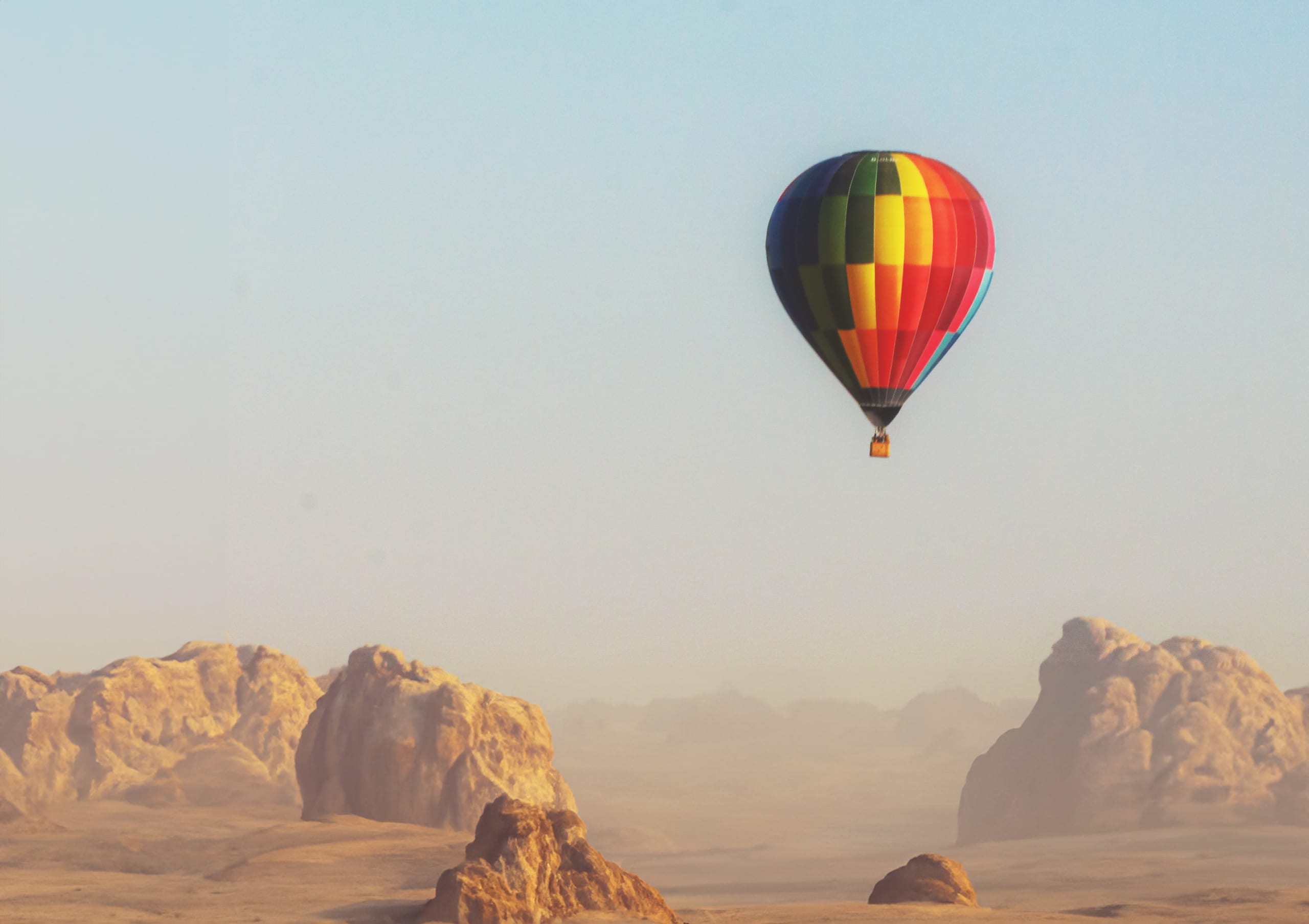 A vibrant multi-colored hot air balloon floats in a pale sky above rugged rock formations and a desert landscape under soft daylight.