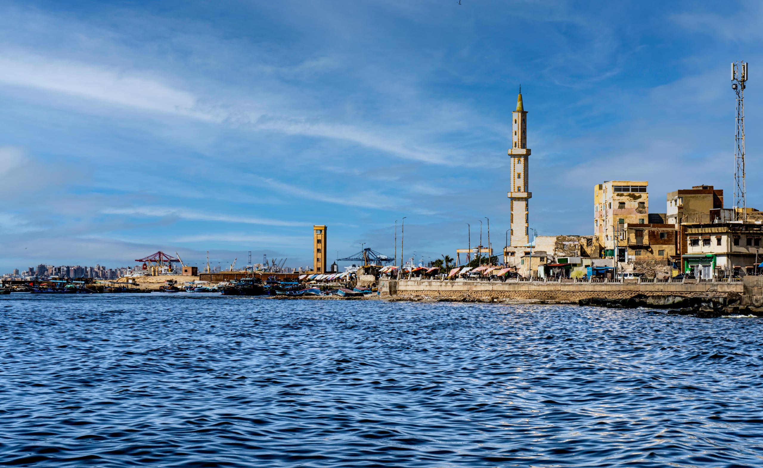 Wide exterior view of a coastal cityscape featuring a prominent tall stone minaret next to older buildings along a seawall, with choppy dark blue water in the foreground and a bright blue sky overhead.