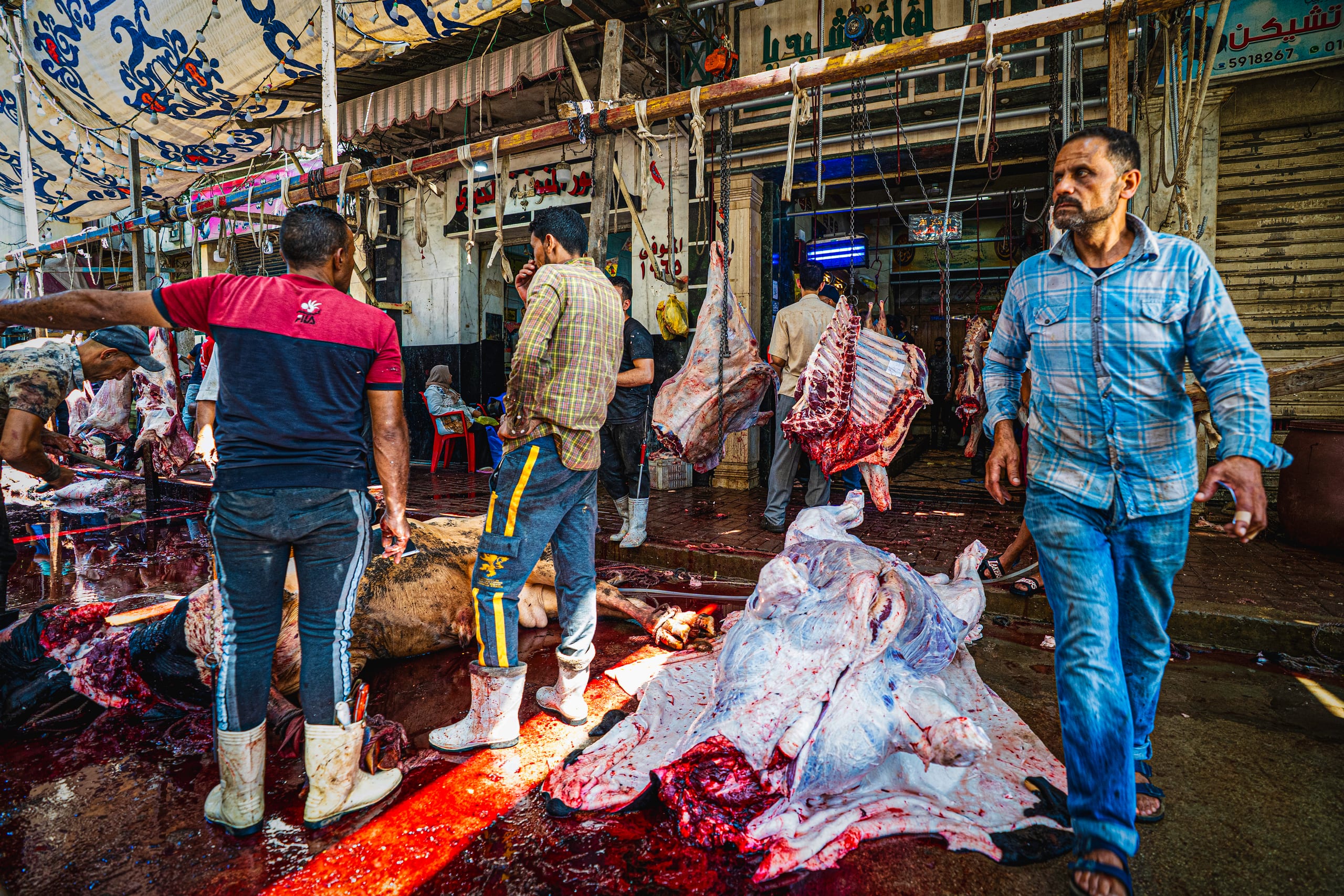 Wide-angle street scene of men working in a busy marketplace next to hanging and skinned animal carcasses on a wet floor, with a man in a blue plaid shirt walking in the right foreground.