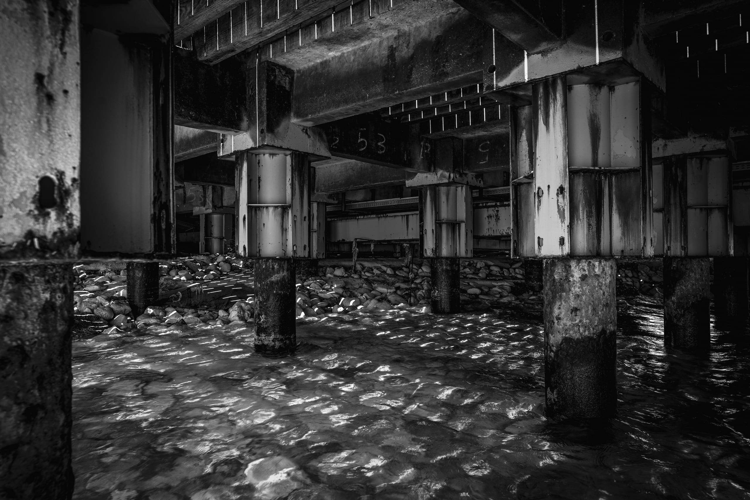 Black and white photograph looking beneath a concrete pier or dock structure, showing heavy support pillars rising from water and a stony shore, with bright sunlight rippling across the surface below.