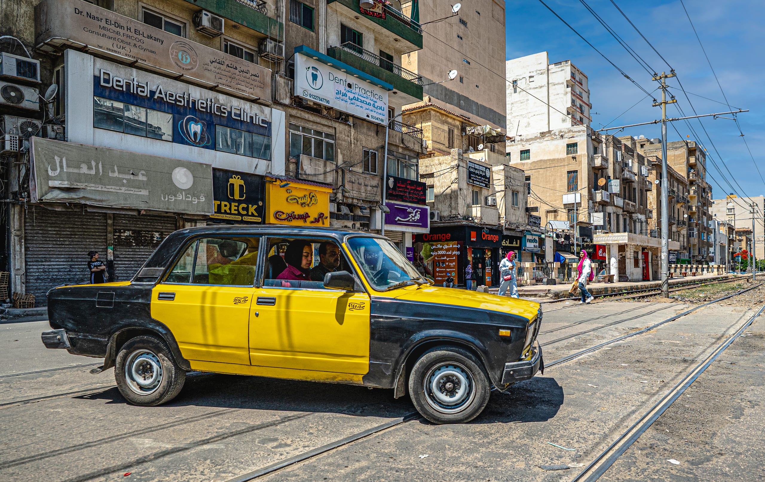A weathered yellow and black sedan moves across street-level tram tracks in front of older multistory commercial buildings with storefront signs and visible pedestrians on the sidewalk.