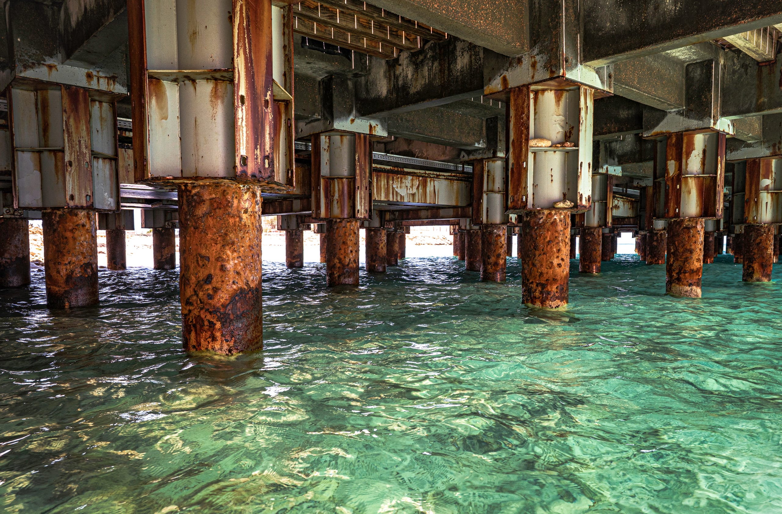 A low-angle shot underneath a concrete and steel pier, showing weathered rust-stained vertical pilings rising out of vibrant turquoise and green seawater.