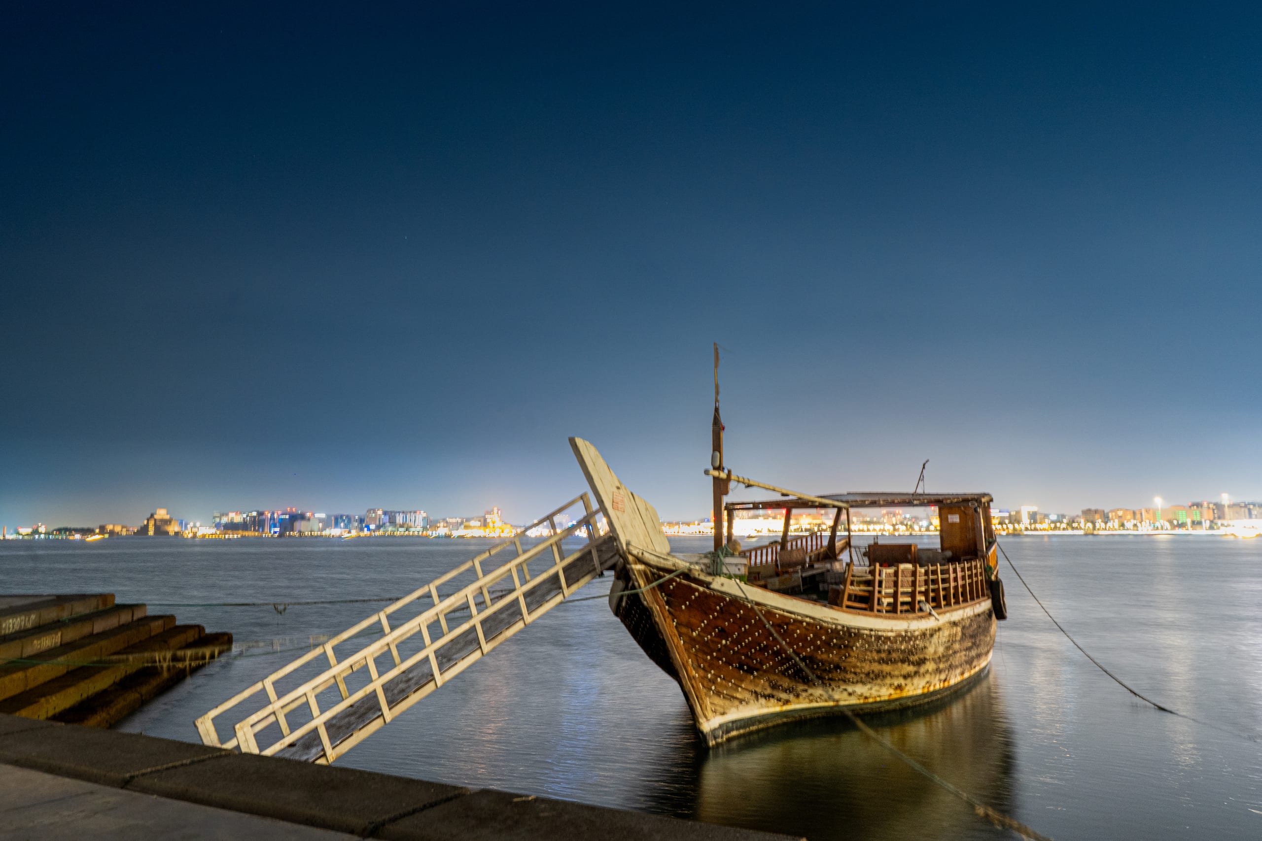 A weathered wooden boat with a lowered gangway is moored by a stone wall on calm water at twilight, with a distant city skyline glowing softly on the horizon under a deep blue sky.