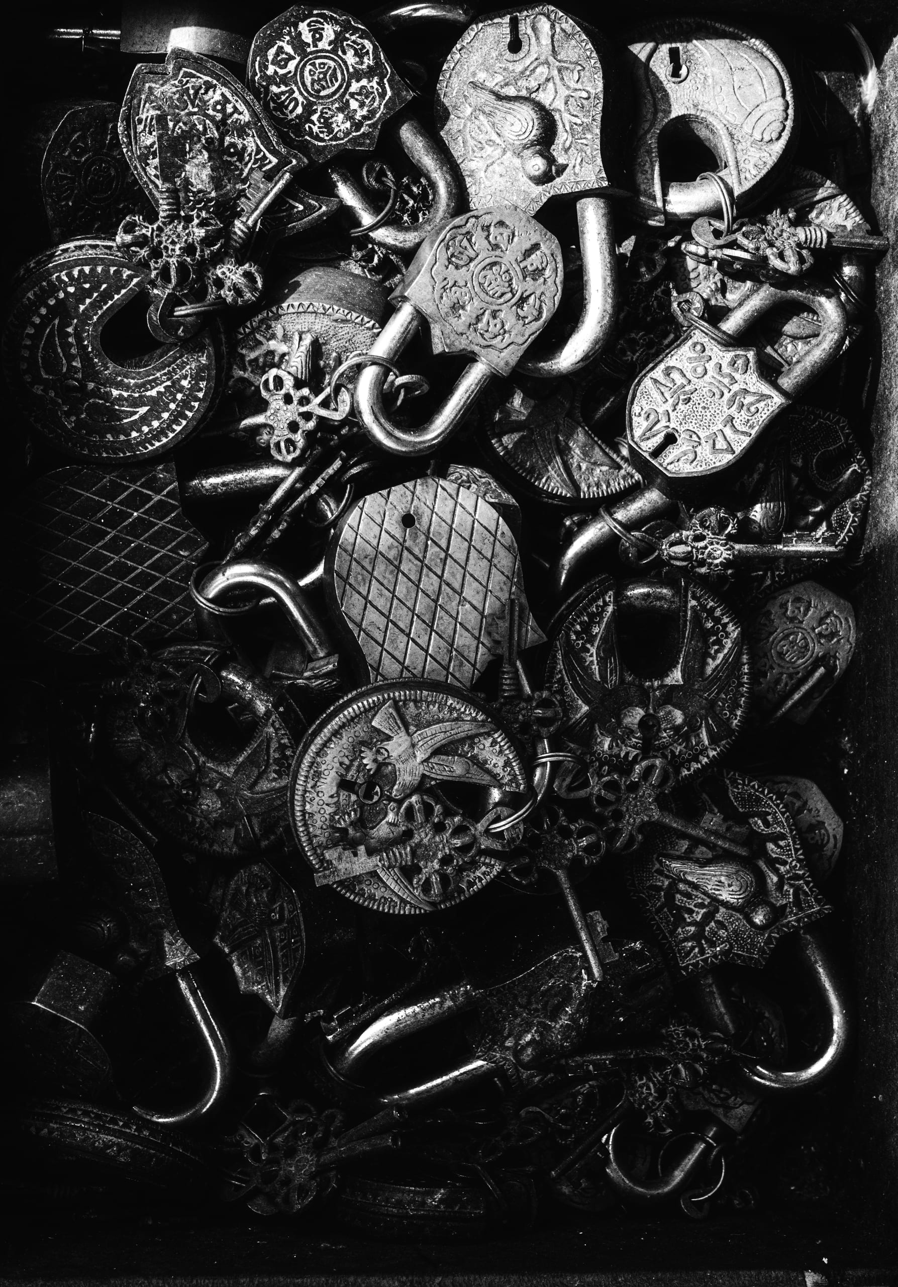 Overhead monochrome close-up of a dense pile of ornate metal padlocks and keys, featuring intricate engravings, metallic highlights, and contrasting deep shadows.