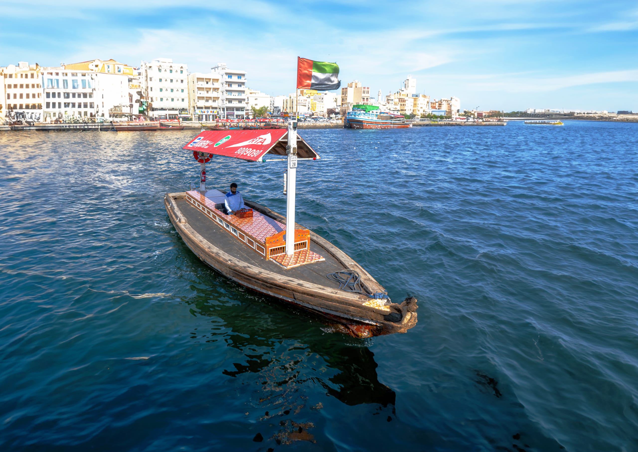 A traditional wooden abra boat with a red-and-white canopy travels across rippling blue water in Dubai. An operator sits mid-boat beneath a flag pole bearing the UAE flag, with low-rise white and cream buildings lining the distant shore.