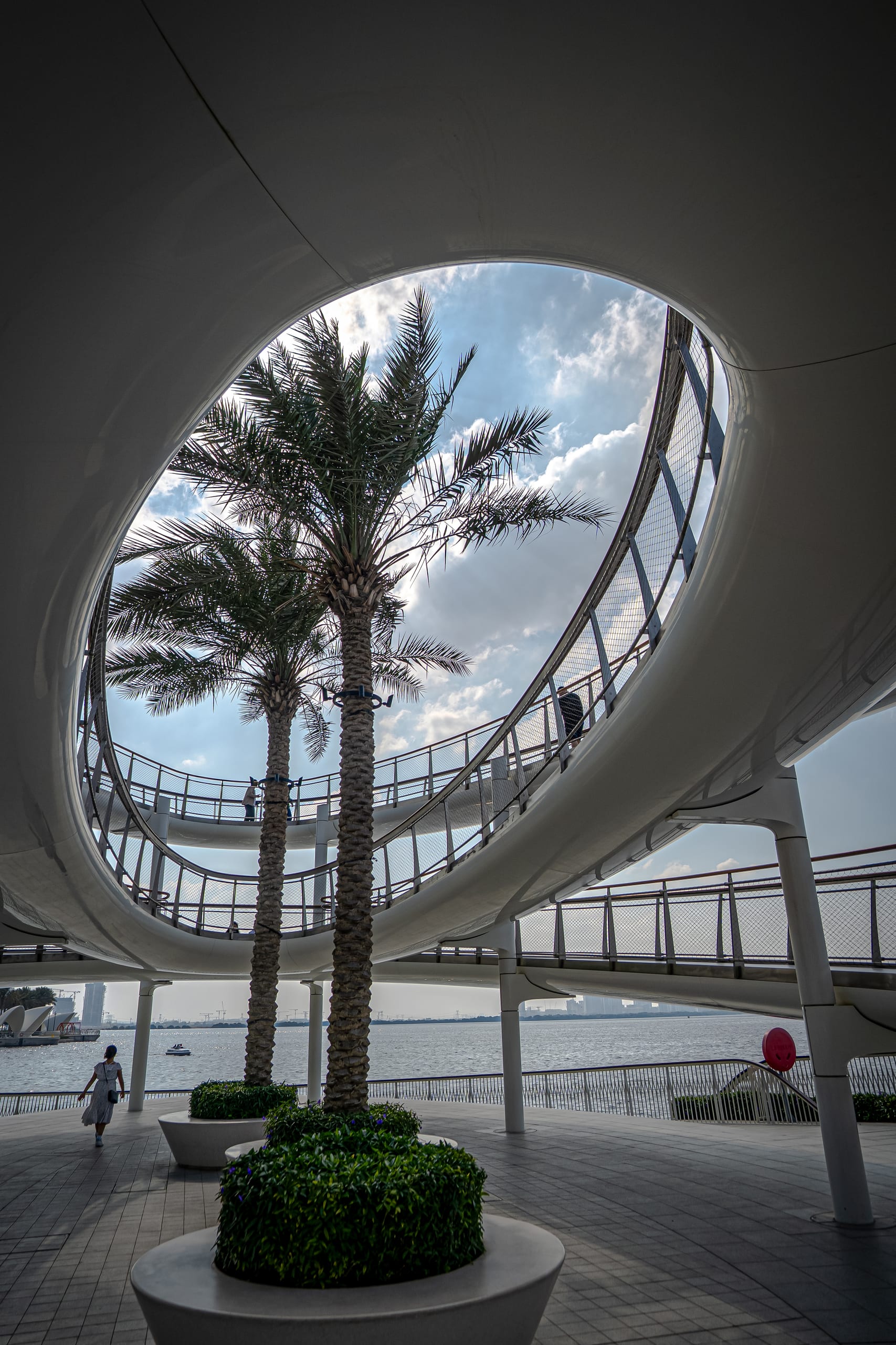 A low-angle view looking upward through a large oval cutout in a white modern architectural structure, revealing two palm trees, a curving elevated walkway with railings, and a body of water in the background.
