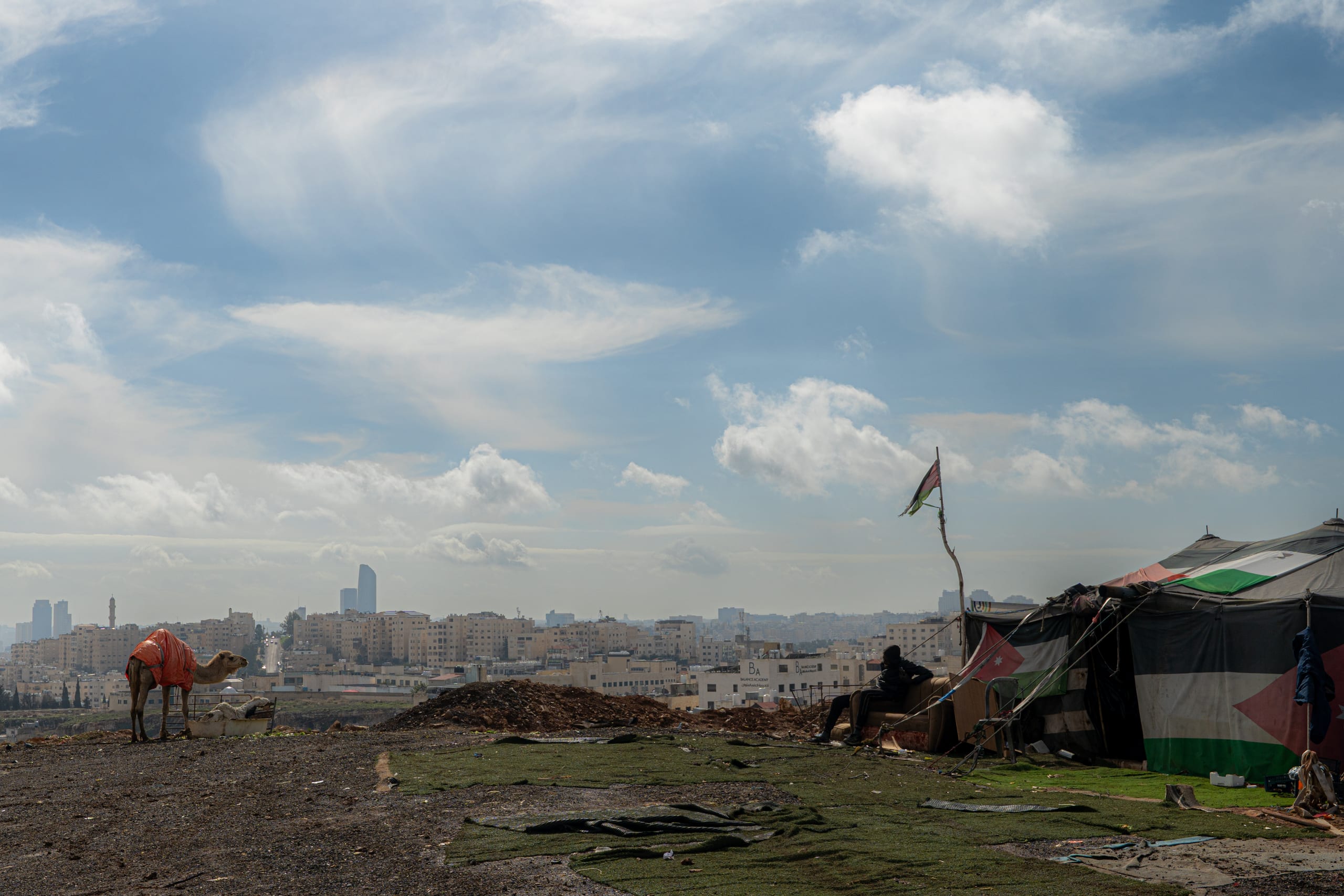Wide-angle outdoor shot showing a camel standing on the left side of a dirt hilltop and a makeshift shelter covered with flags on the right. In the background, a dense city spreads beneath a bright blue sky filled with scattered white clouds.
