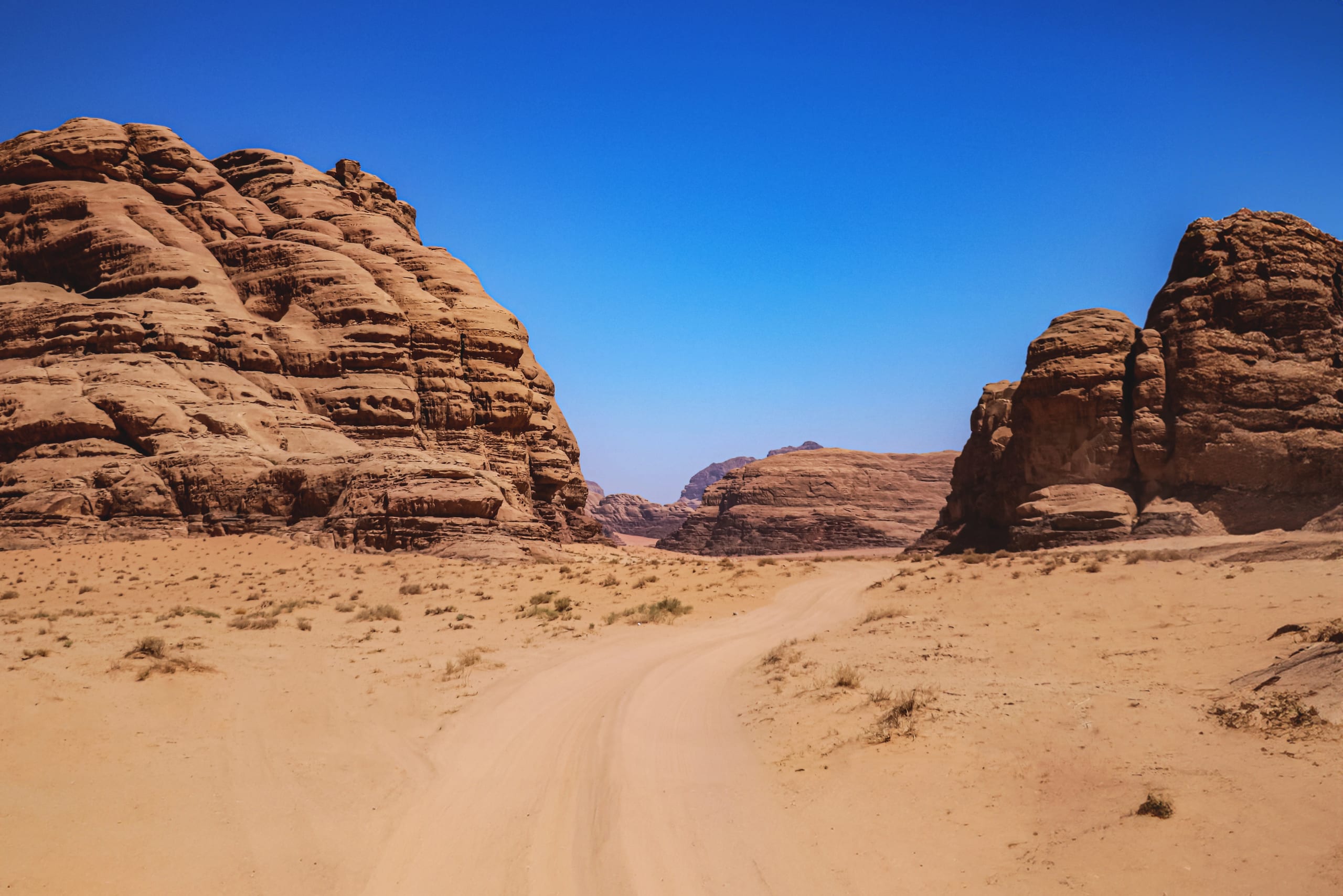 A wide angle landscape shot showing a sandy vehicle track curving through a desert valley between massive eroded rock formations under a clear blue sky.