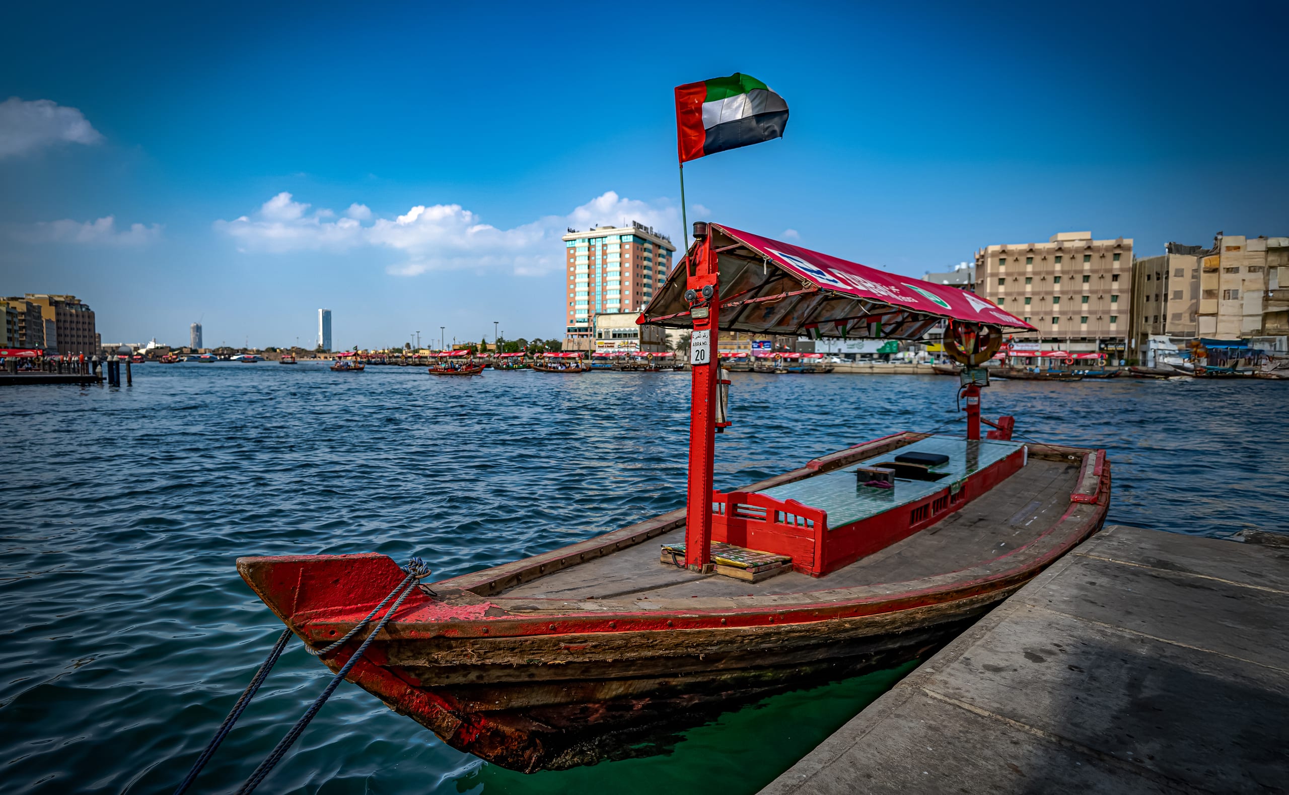 A traditional wooden water taxi with a red canopy and a national flag is tied to a concrete dock, floating on deep blue water with buildings visible across the waterway.