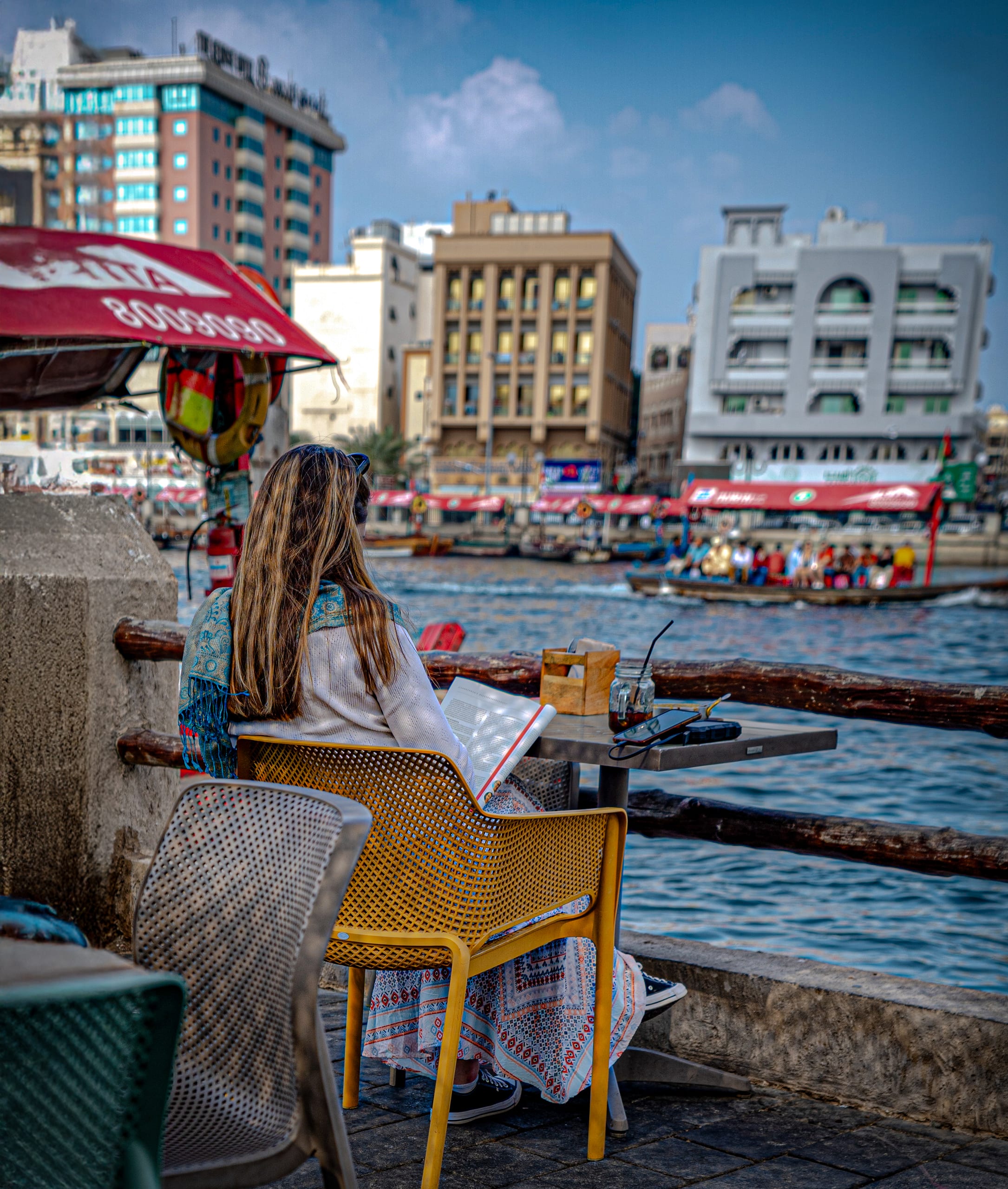 A woman with long blonde hair seen from behind, sitting in a yellow chair at a wooden outdoor table by the water, reading a large open book. Across the water, classic wooden boats carry passengers past a cityscape of traditional and modern buildings under a bright sky.