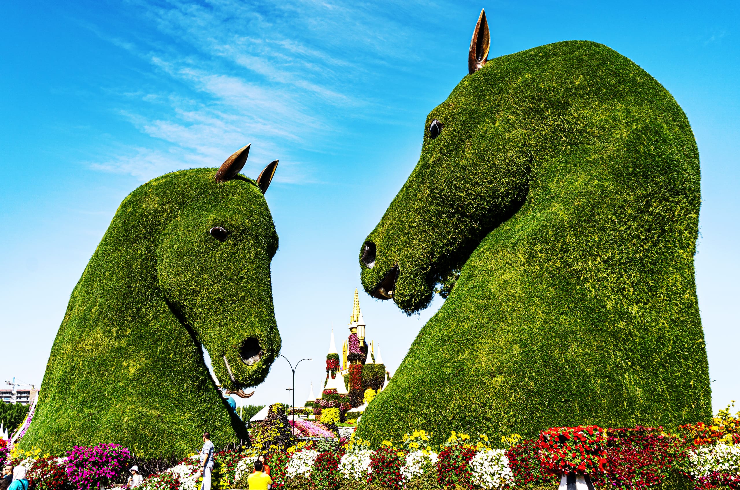 Low-angle shot of two giant green topiary horse heads facing each other under a clear blue sky, with colorful flower beds lining the foreground and a small castle structure visible in the gap between them.
