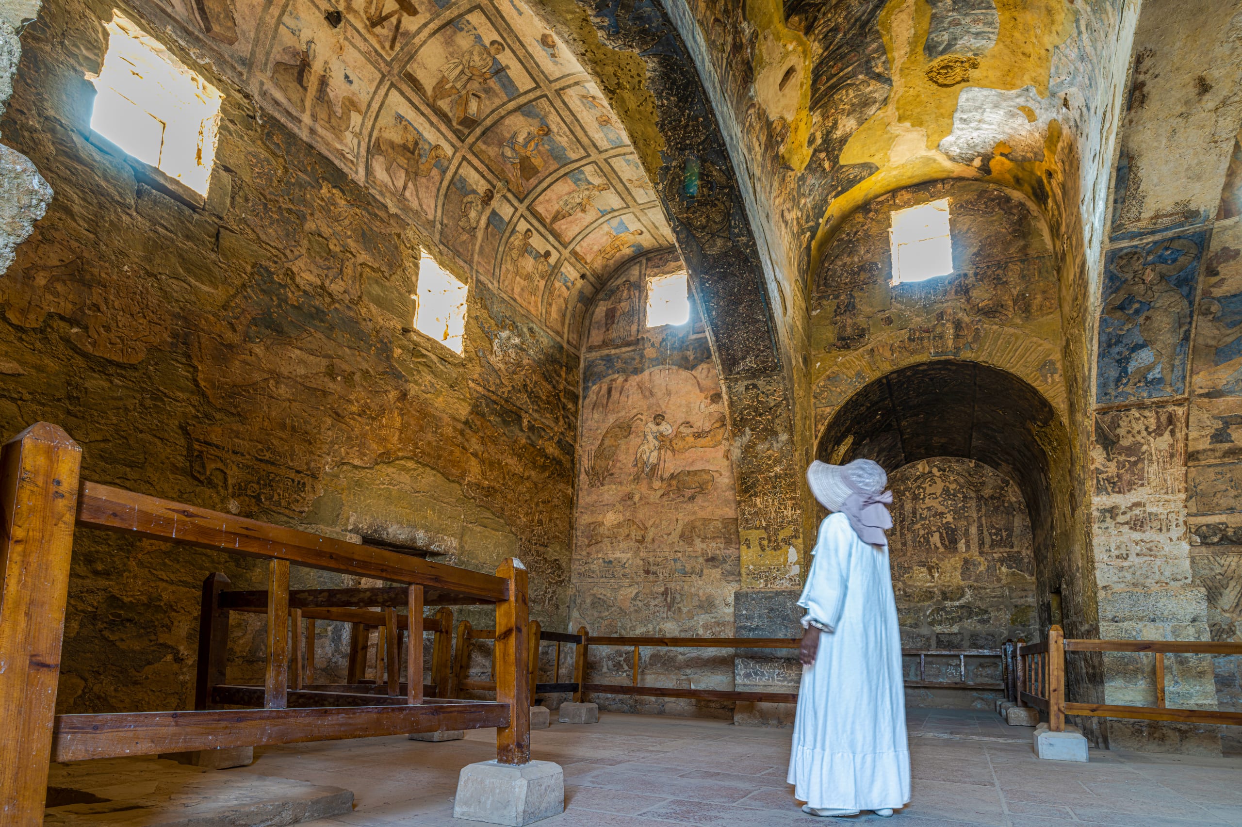 A person wearing a white dress and hat stands inside a vaulted stone chamber, looking upward at weathered frescoes on the ceiling and walls, framed by low wooden barriers in the foreground.