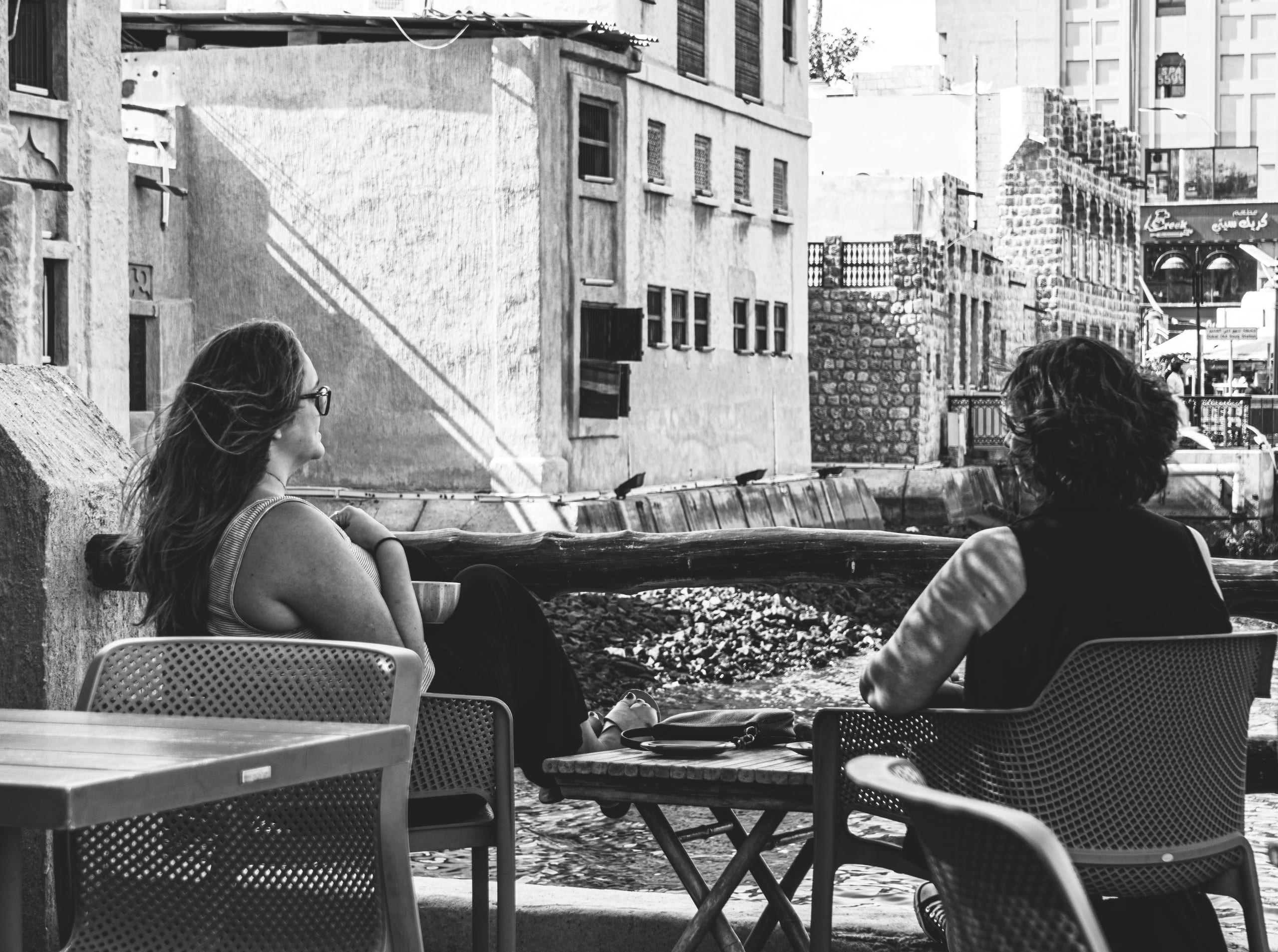 Monochrome street-level view of two women sitting at a table outdoors facing a waterfront, with historic stone buildings and harsh diagonal shadows in the background.