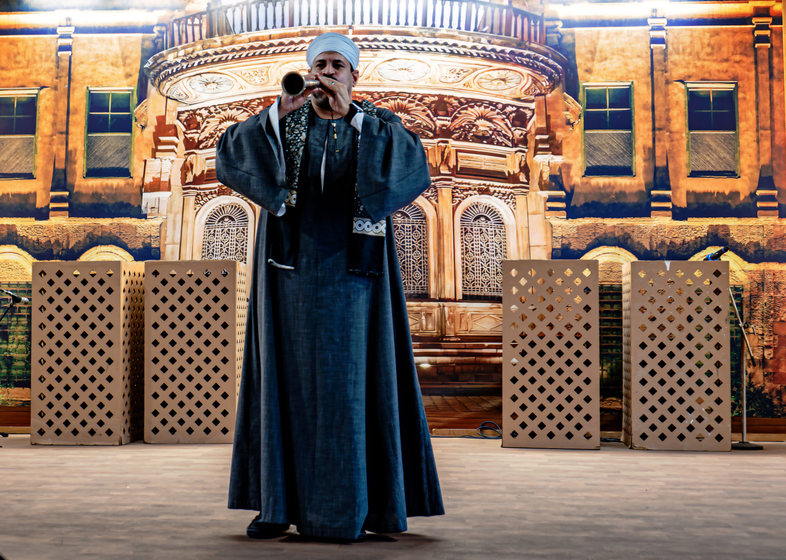 A full-length portrait of a male musician in traditional garments playing a wind instrument on stage, positioned before a painted architectural backdrop and decorative lattice panels.