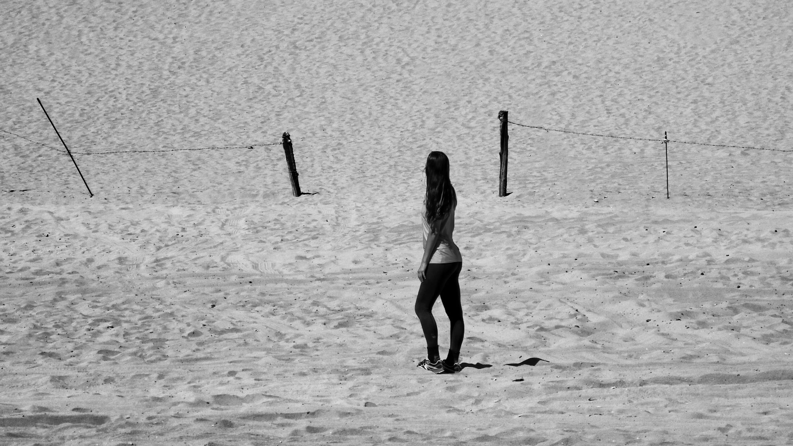 A person with long hair stands in profile on a textured sandy surface, viewed mid-distance with a simple wire fence crossing the upper background in a black-and-white composition.