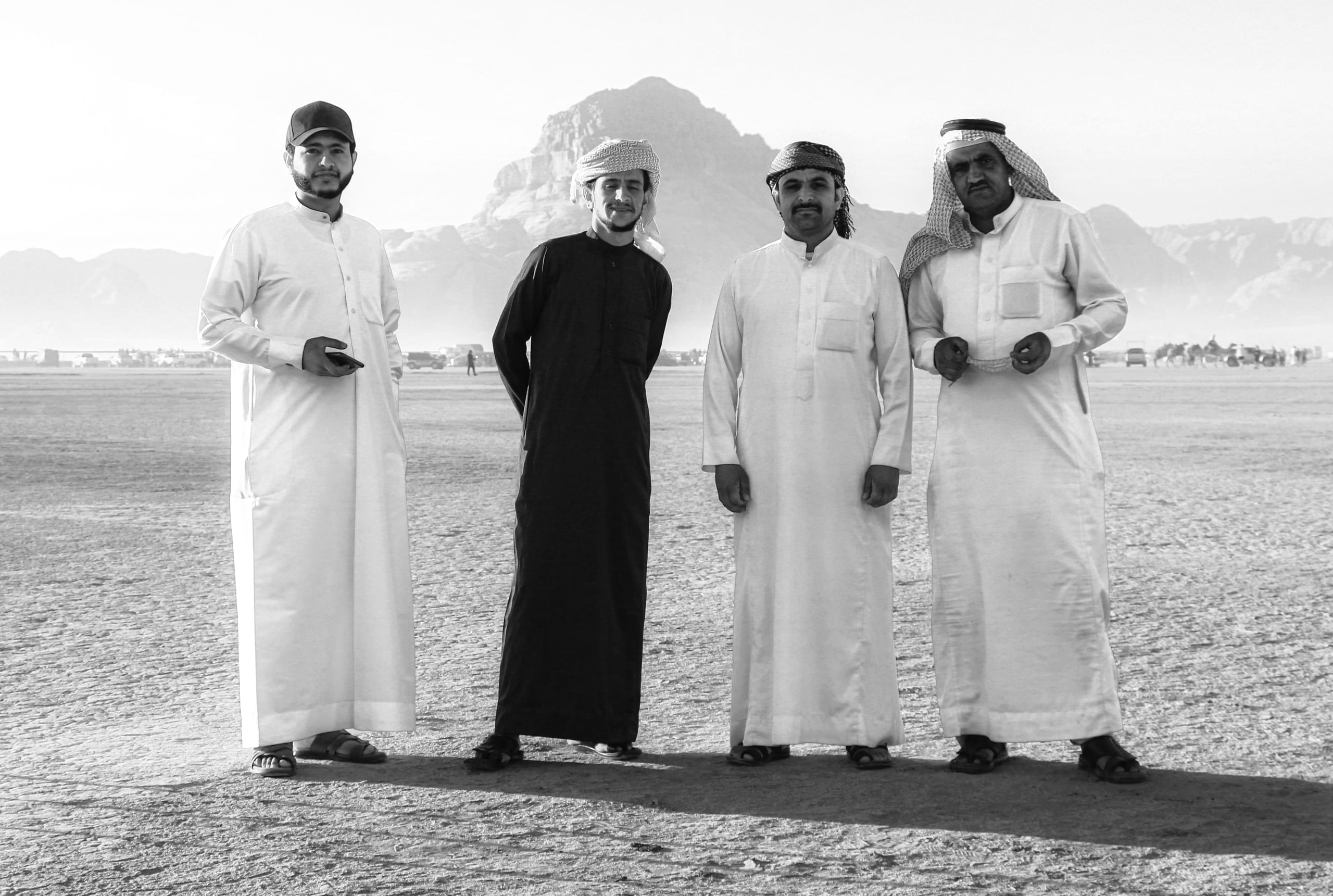 Four men wearing traditional robes standing side by side outdoors in a flat, bright landscape with distant mountains in the background, captured in monochrome.