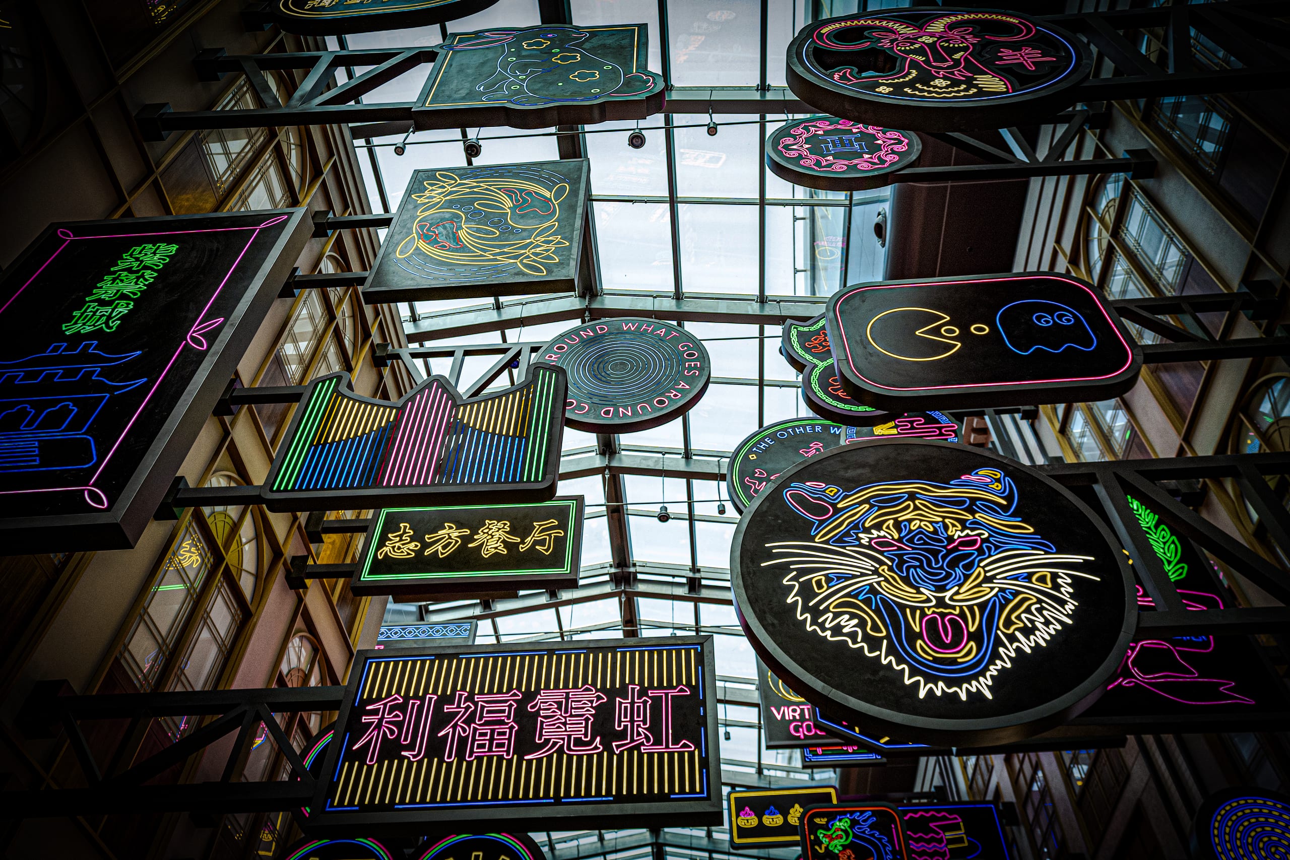 Low-angle view of numerous colorful neon signs hanging from a dark metal grid framework beneath a bright glass atrium roof.