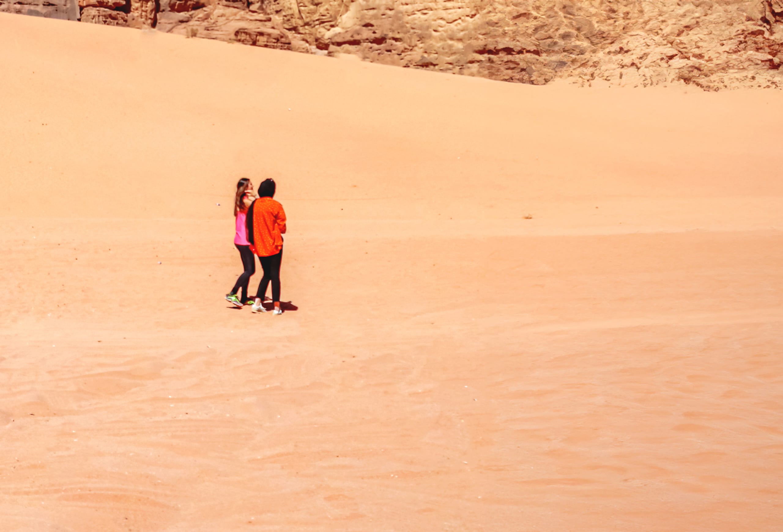 Two people wearing bright orange and pink jackets walking together across a vast orange sand dune with rocky cliffs visible in the background.