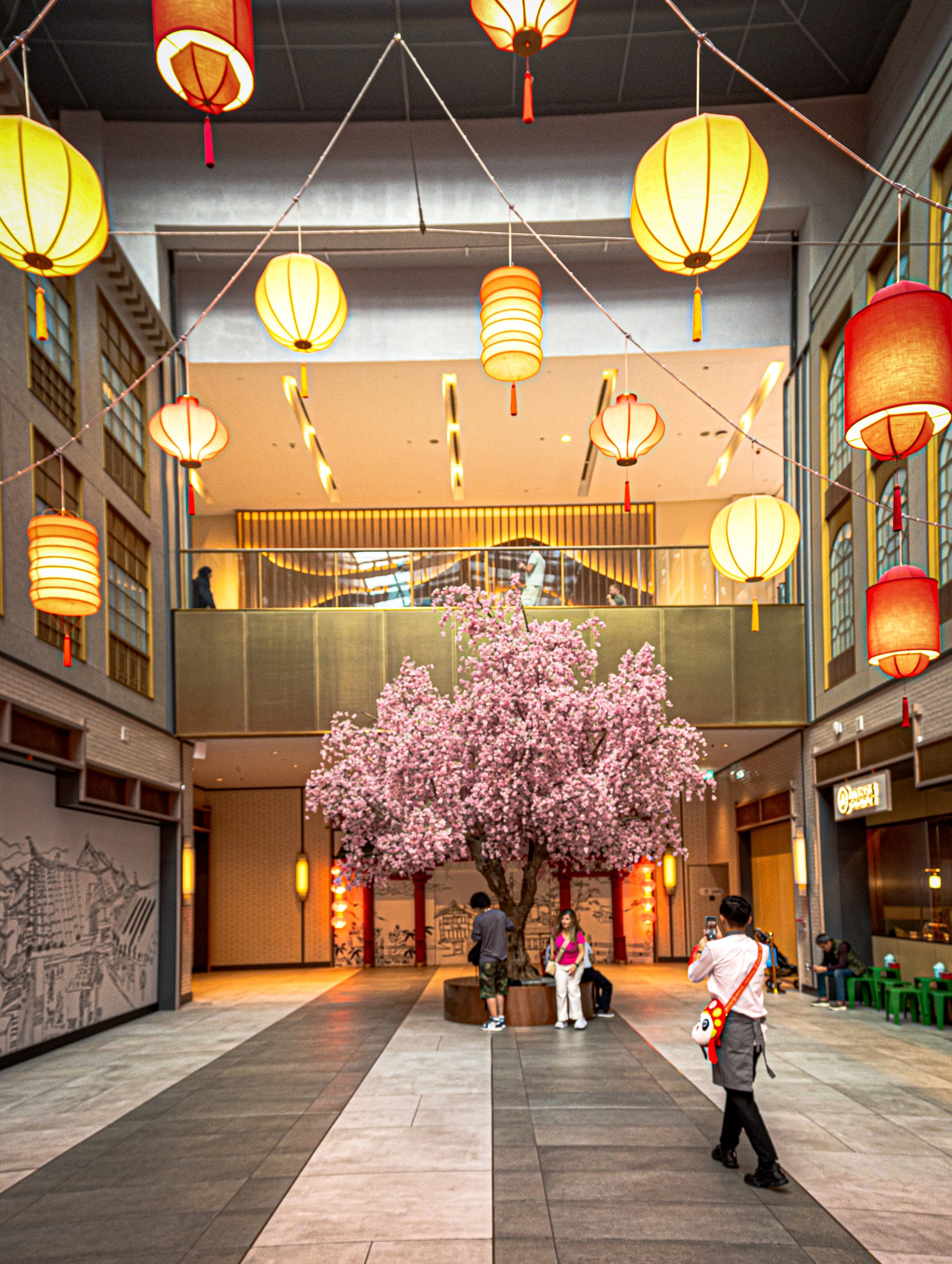 An interior shopping space decorated with dozens of glowing red and yellow paper lanterns suspended on strings across the ceiling above a large blooming pink cherry blossom tree and strolling visitors.