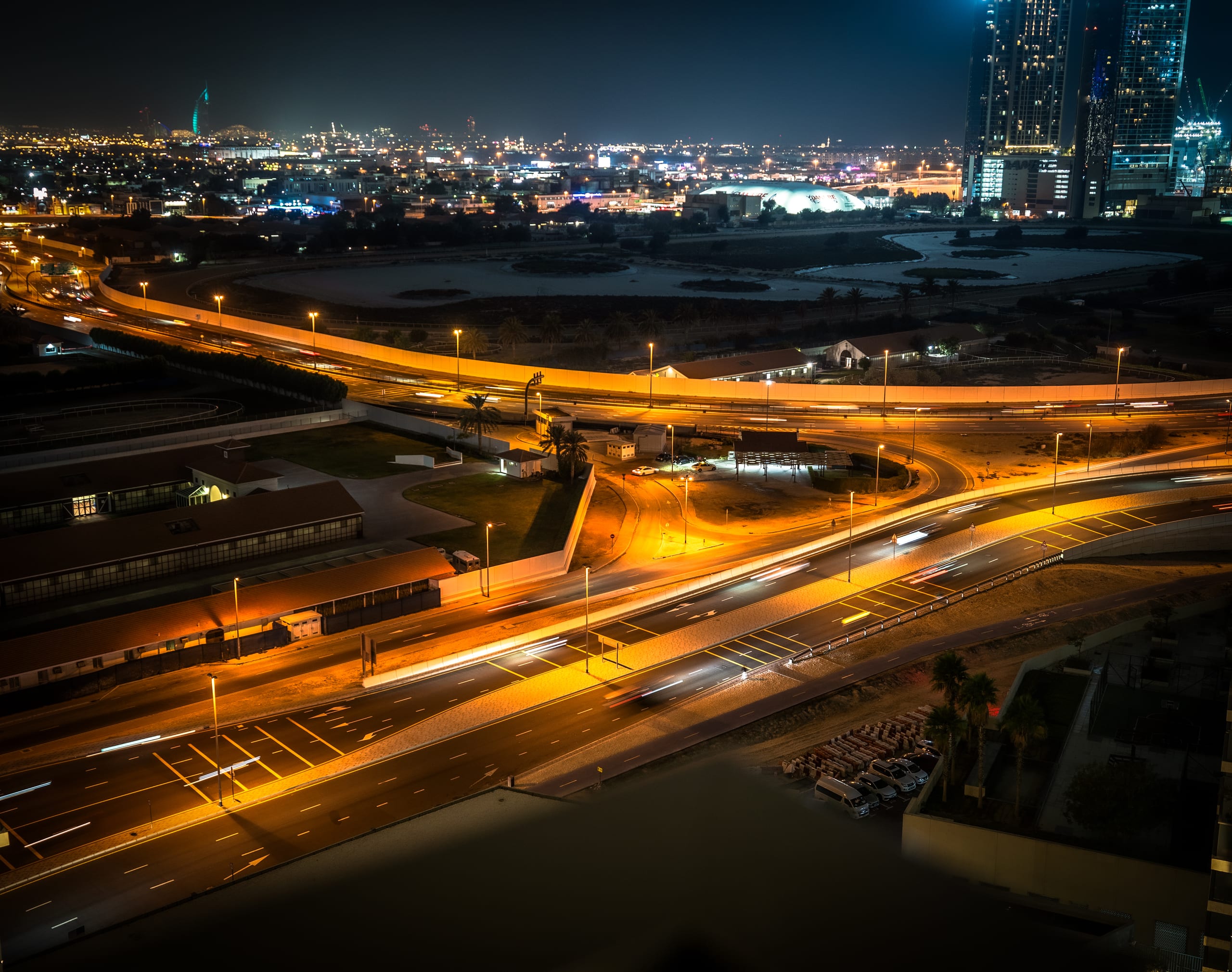 High-angle night shot of illuminated multi-lane roads with long-exposure light trails from vehicles, set against a dark city skyline in Dubai, UAE.