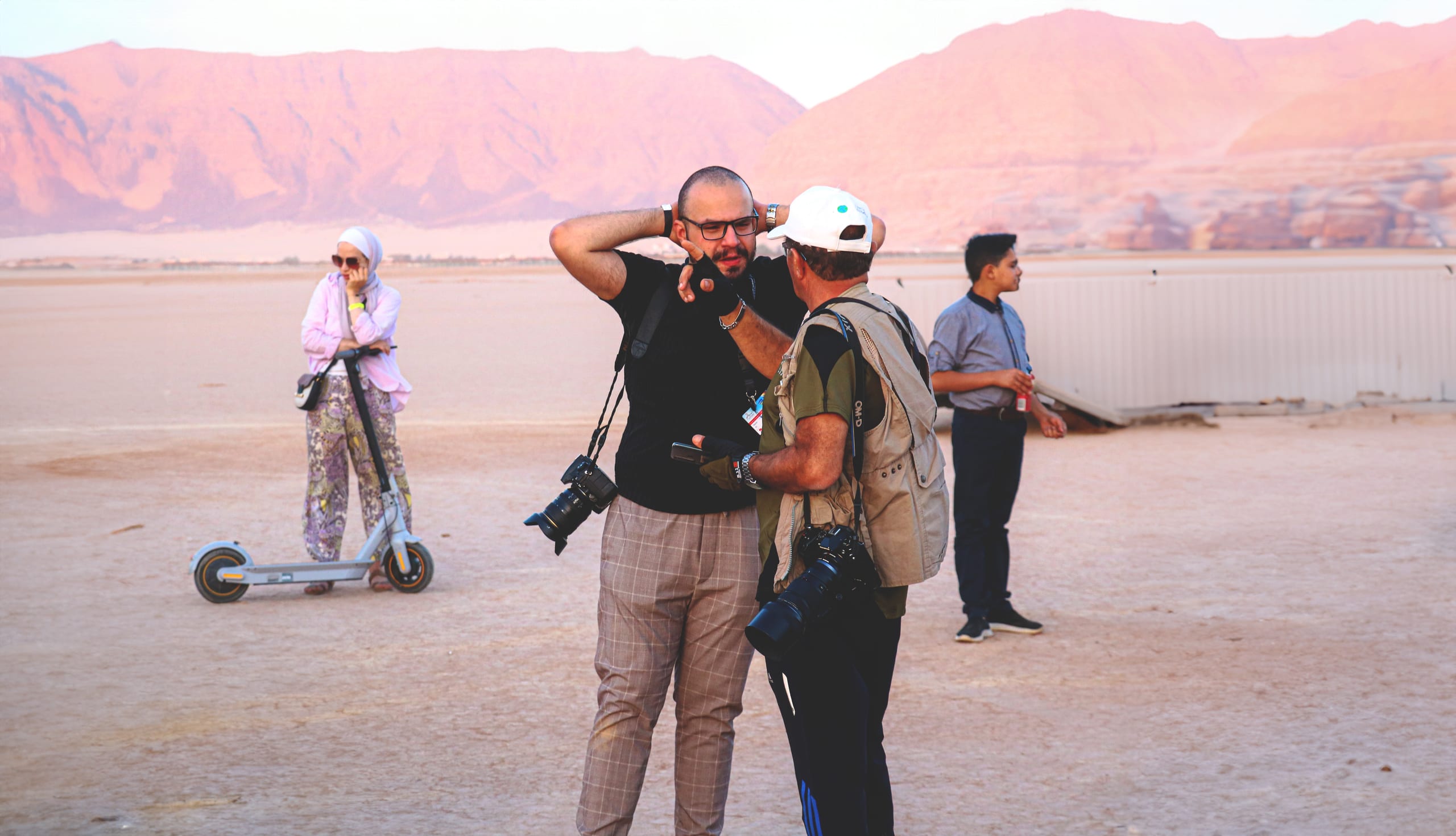 Two men with cameras standing and talking in a dry, open landscape with mountains in the background, flanked by a person on an electric scooter to the left and another person standing to the right.