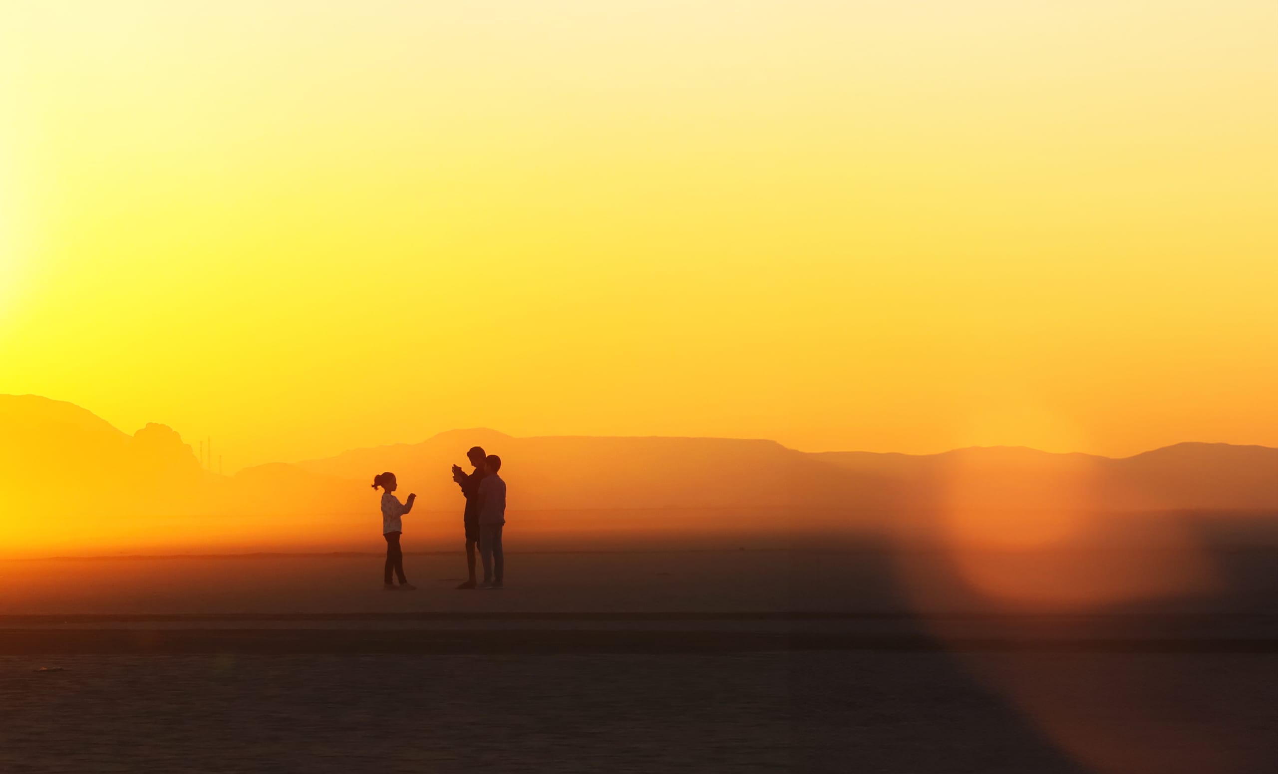 Silhouette of three figures standing against a bright yellow and orange desert sky with distant mountains on the horizon.