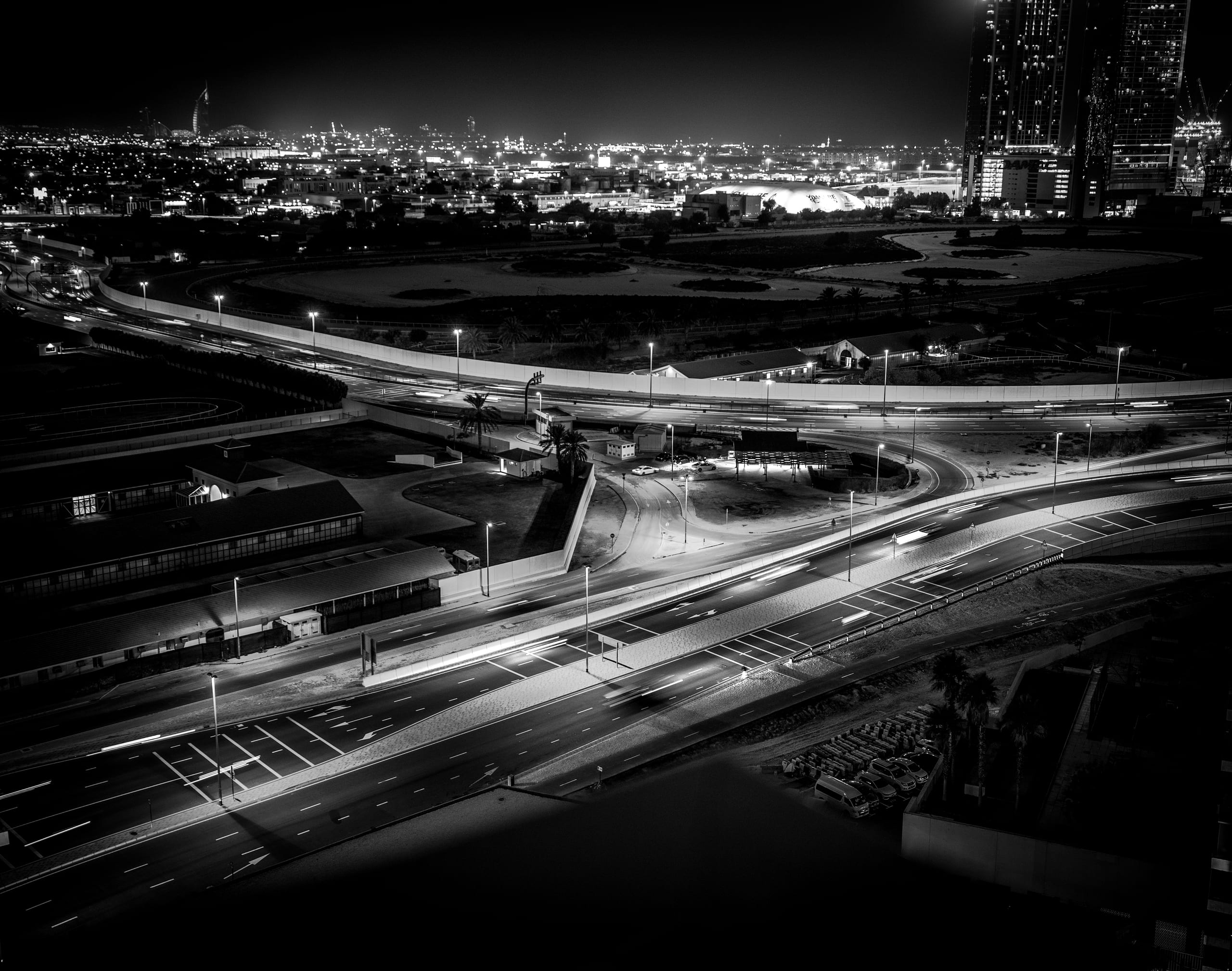 High-angle black and white night shot of urban roads with long-exposure light trails from cars, buildings, and distant city lights.
