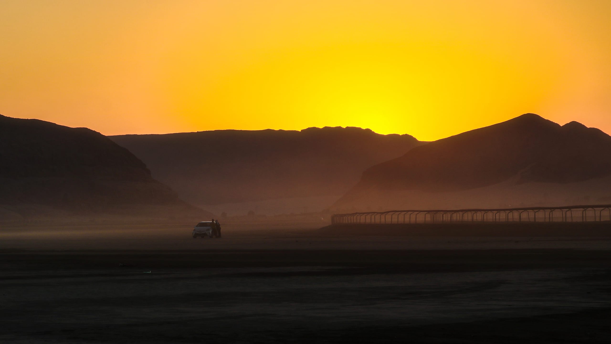 A wide-angle desert landscape at sunset, featuring a glowing yellow sky above dark silhouette mountains, a hazy valley floor, and a small vehicle on the left side near a fence line.