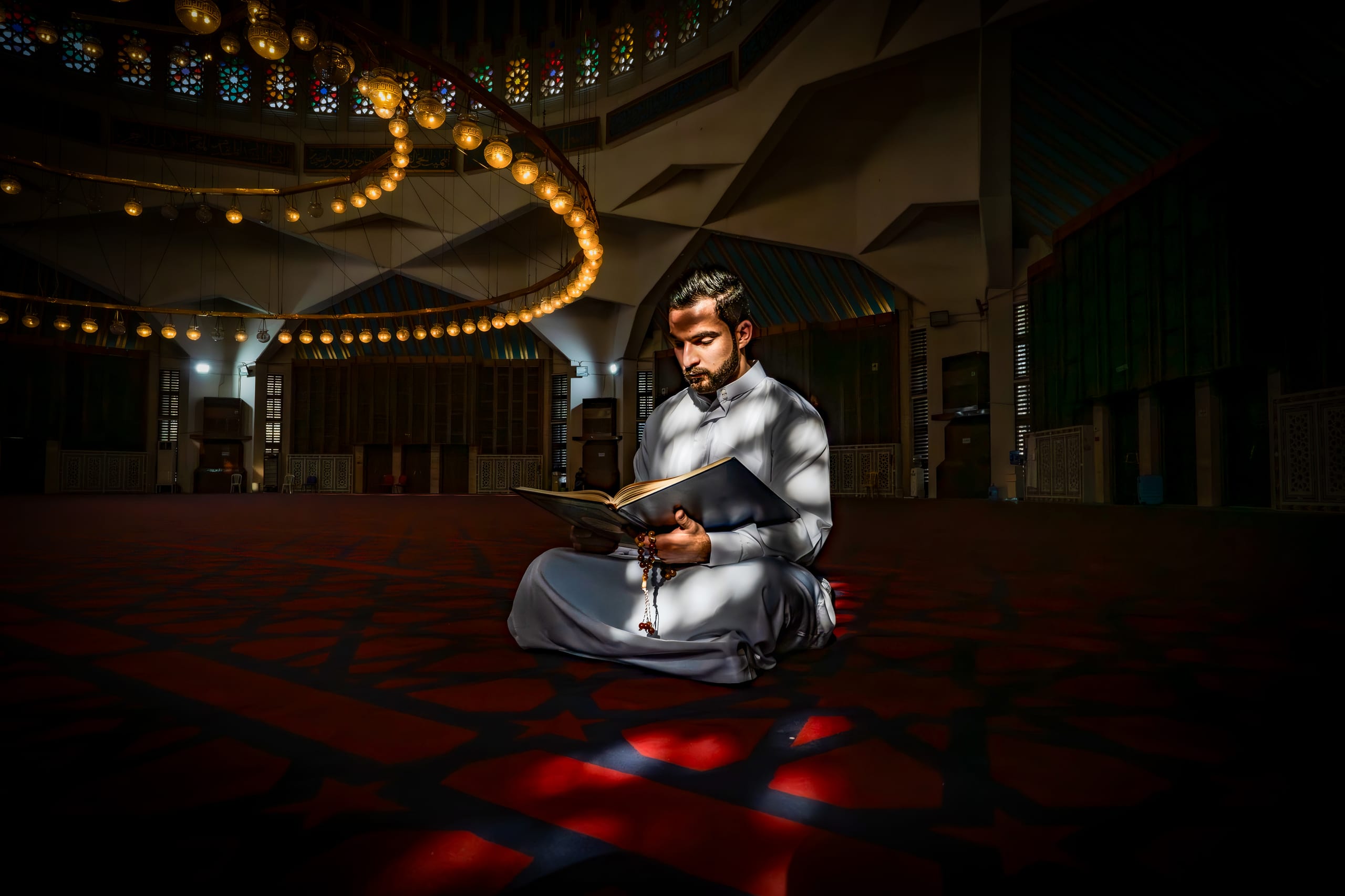 A man in traditional white attire sits cross-legged on a patterned red carpet in a large dimly lit hall, reading an open book while illuminated by a beam of sunlight. A large illuminated circular chandelier curves across the upper left ceiling.
