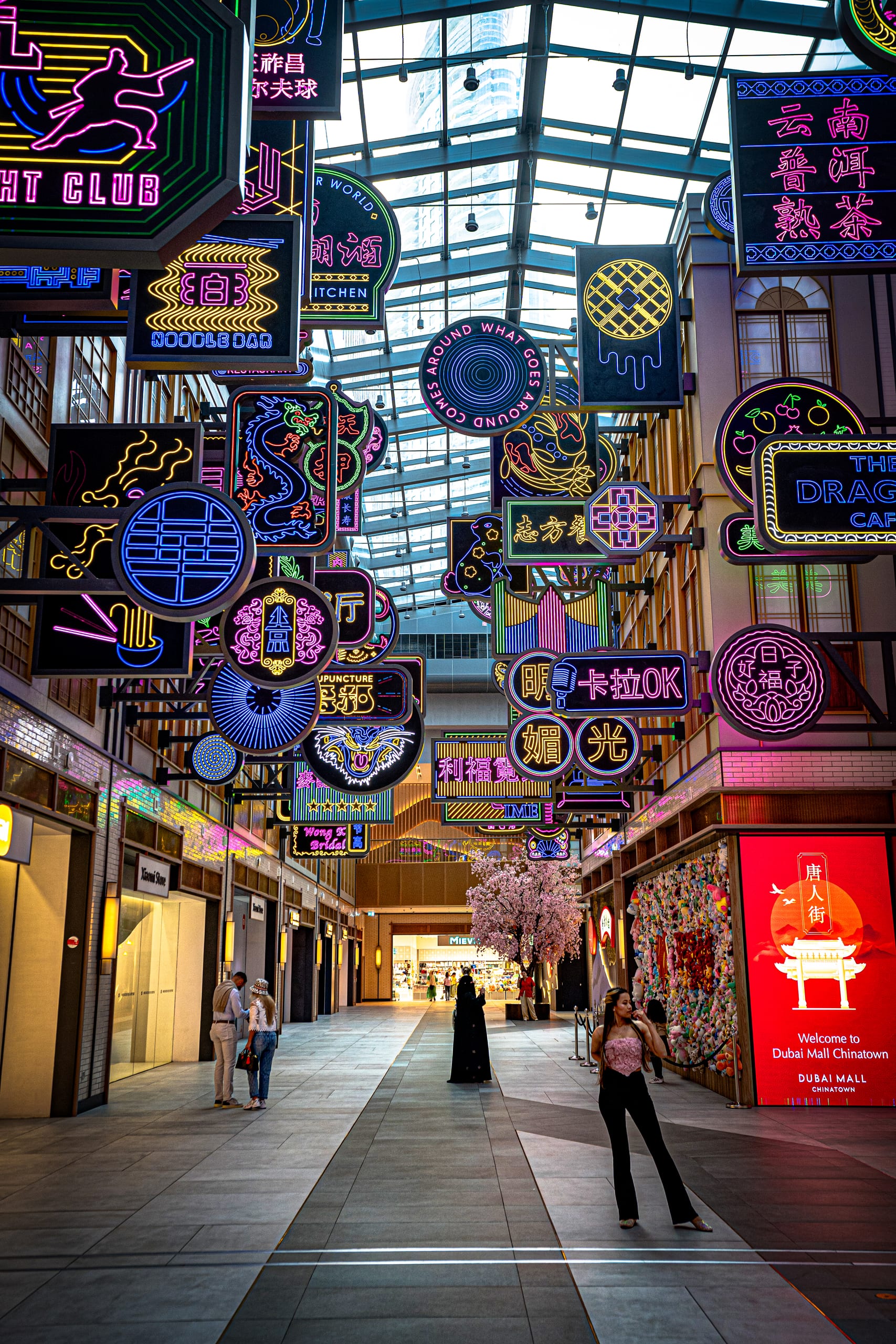 Wide-angle view of a covered pedestrian street or mall corridor crowded with suspended glowing neon signs in various shapes, colors, and Chinese characters, with pedestrians walking along the tiled floor below.