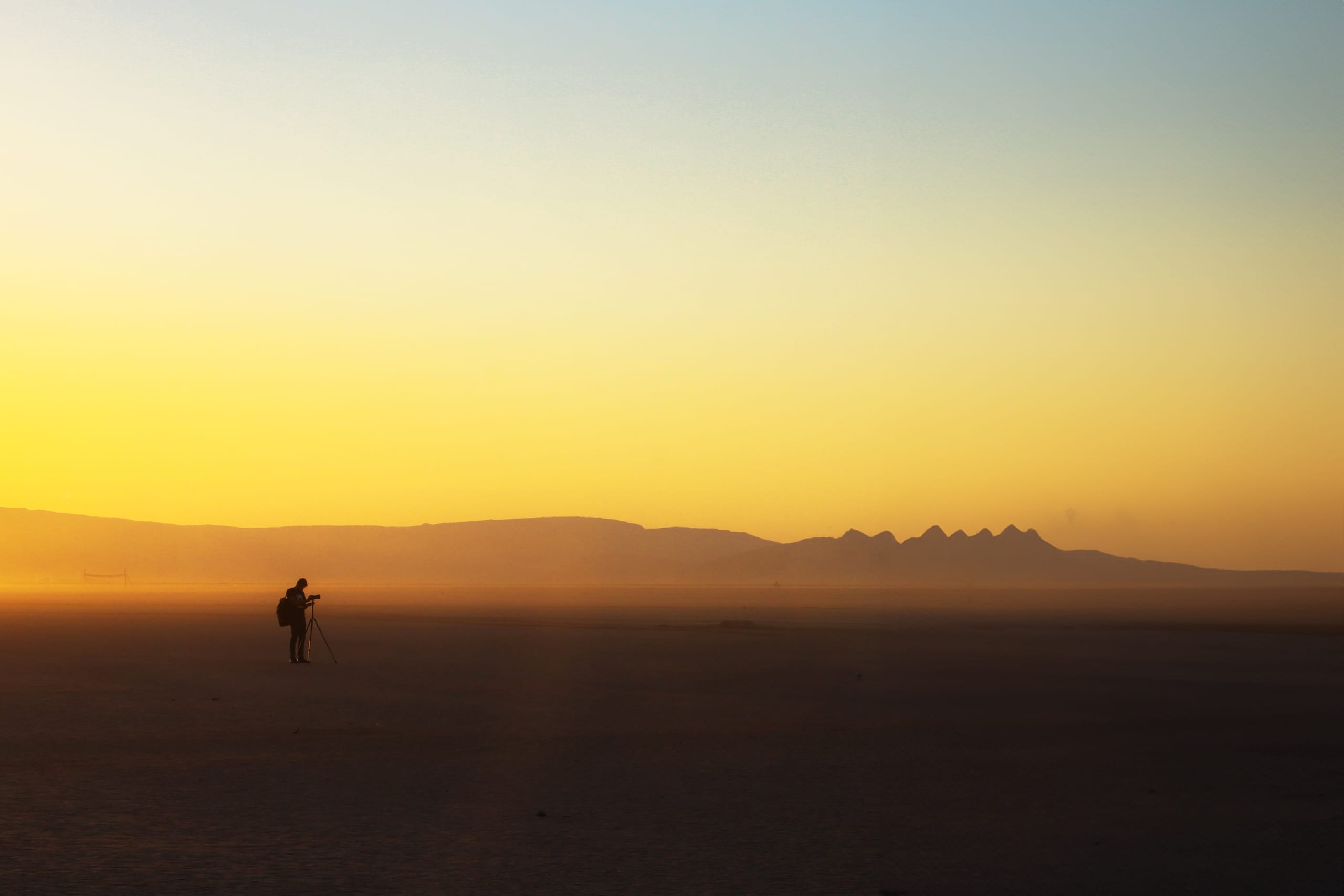 Wide-angle landscape shot of a silhouetted photographer with a tripod in a flat desert beneath a bright yellow and blue twilight sky, with distant mountains along the horizon.
