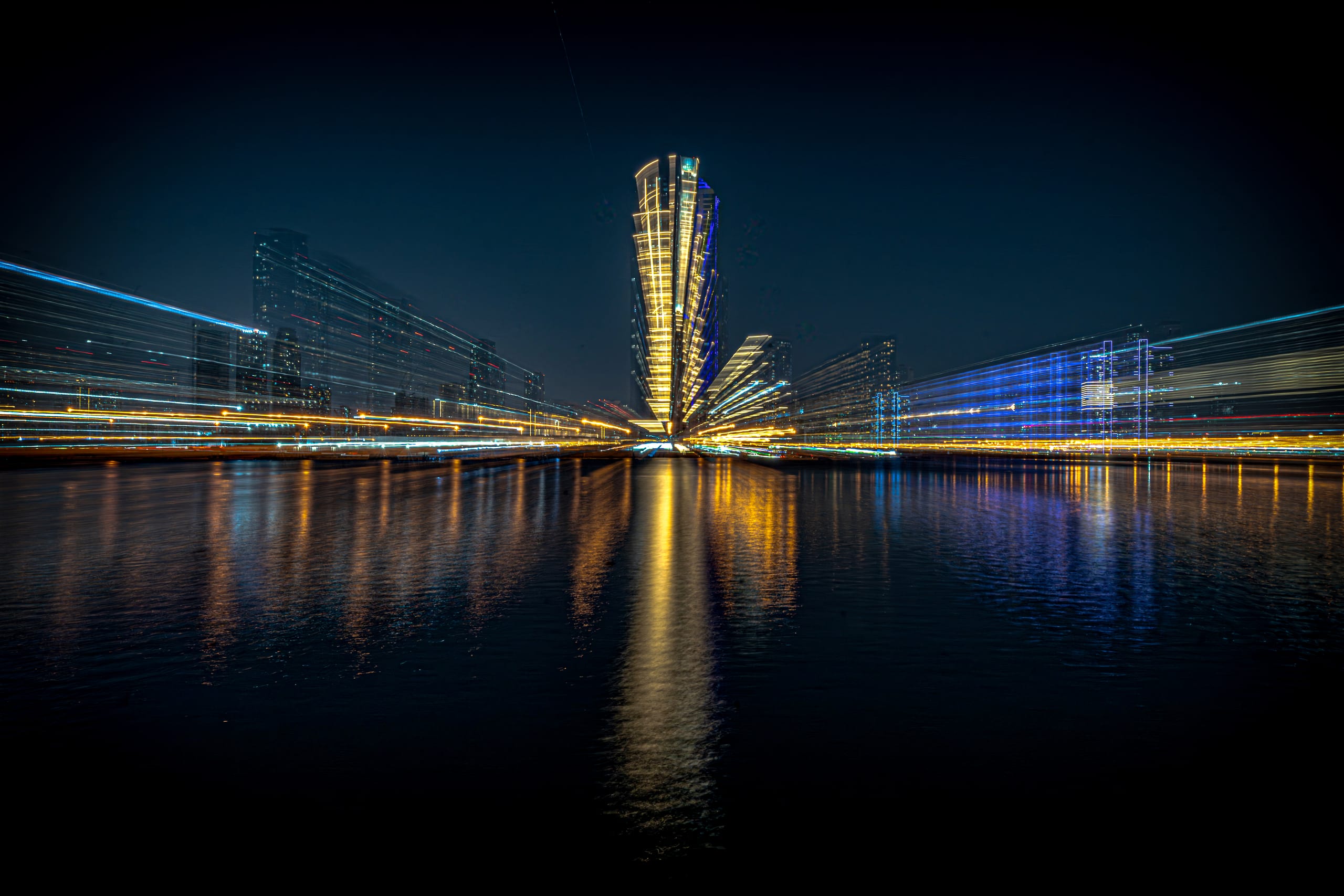 Night long-exposure photograph showing streaking golden and blue city lights radiating outward from a central illuminated skyscraper, reflecting onto dark water below.