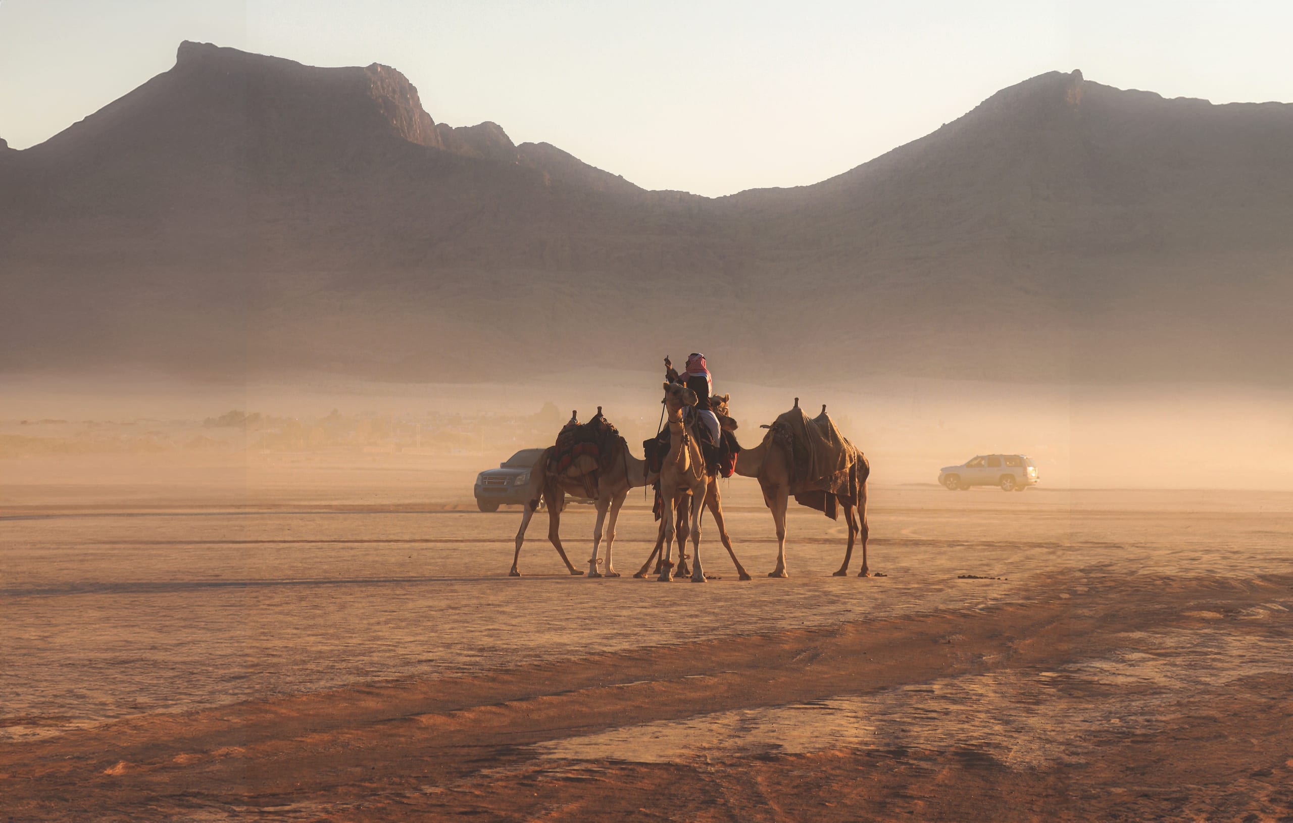 A wide shot of three saddled camels carrying a rider in a dusty, sunlit desert plain, framed by distant rugged mountains and a low hazy horizon.
