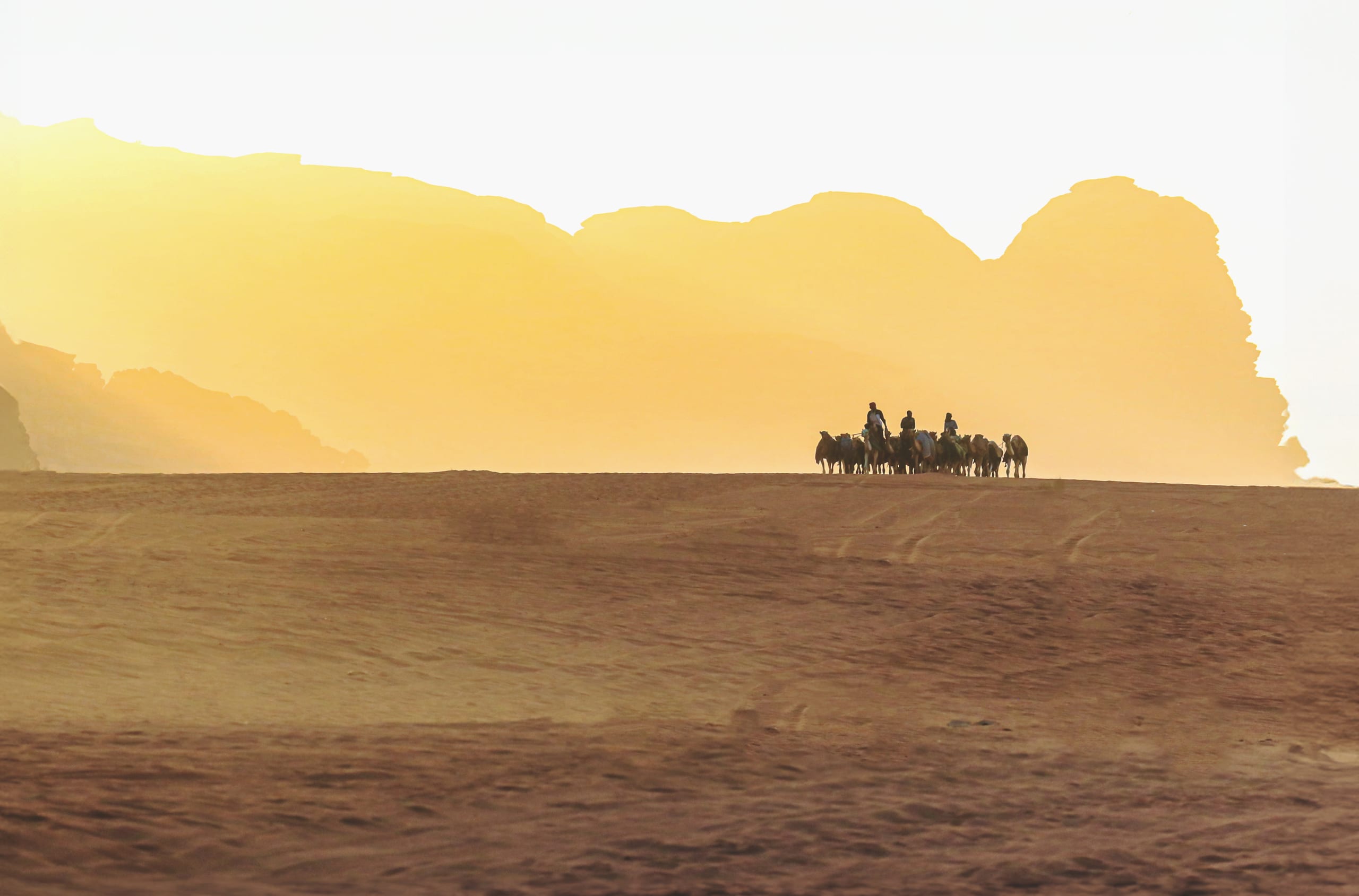 A group of people riding camels in a silhouetted caravan across a golden desert dune under bright backlit sky, with large mountain formations in the background.