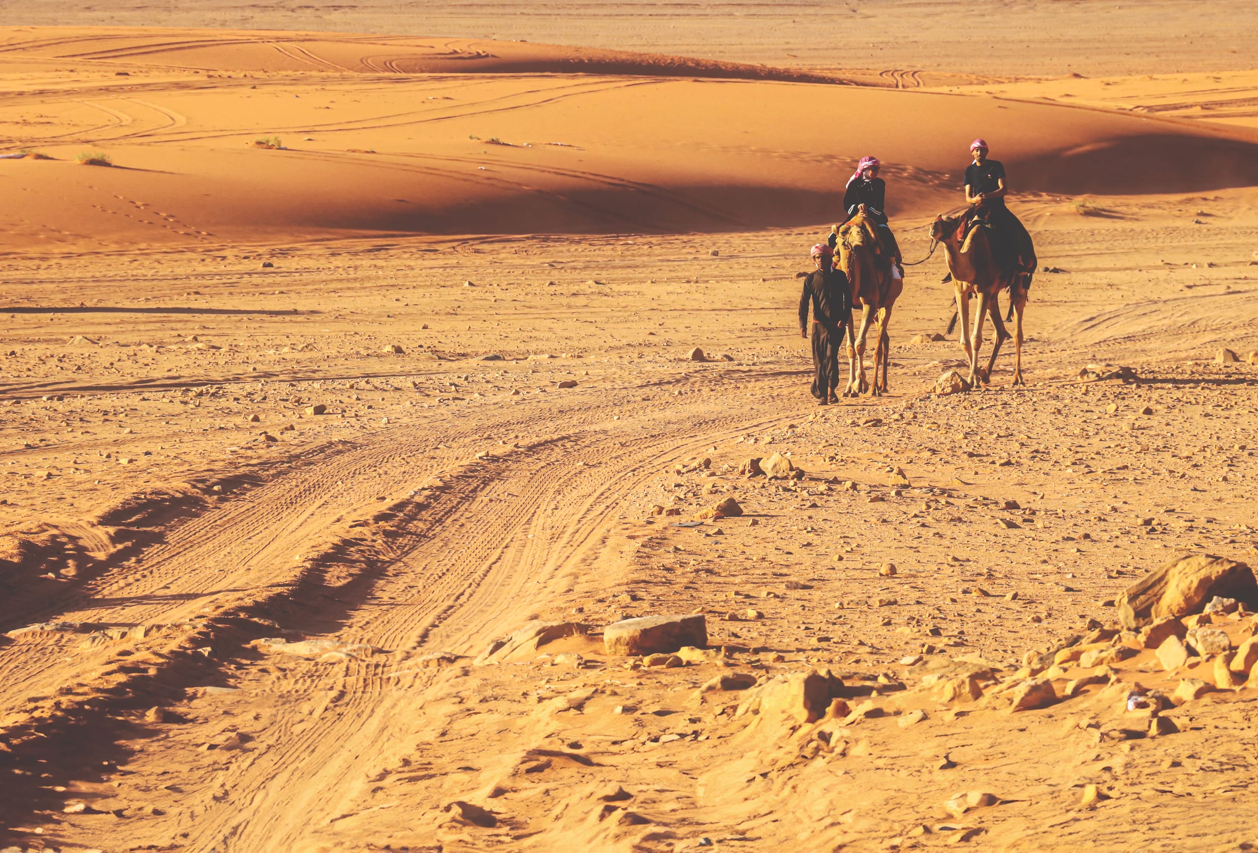 Wide-angle outdoor shot of two people riding camels and one person walking across a vast sand and rock desert landscape under warm sunlight.