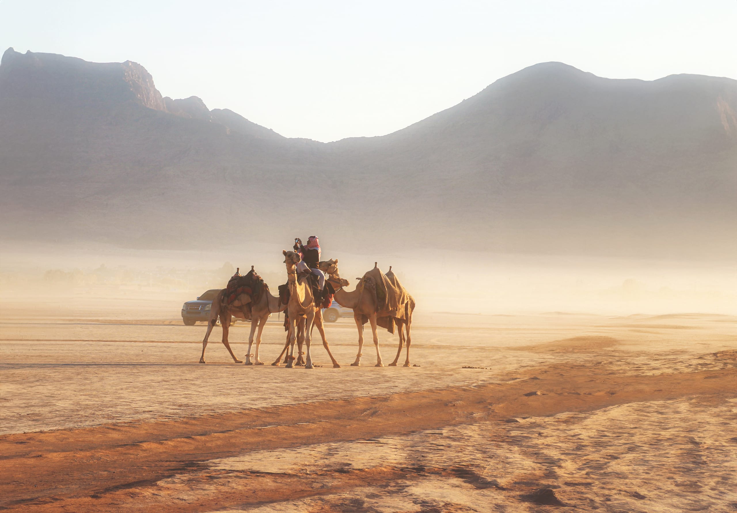 Wide-angle landscape view of three saddled camels with riders standing together in a sandy desert plain, surrounded by a heavy morning haze and large mountains under a bright sky.