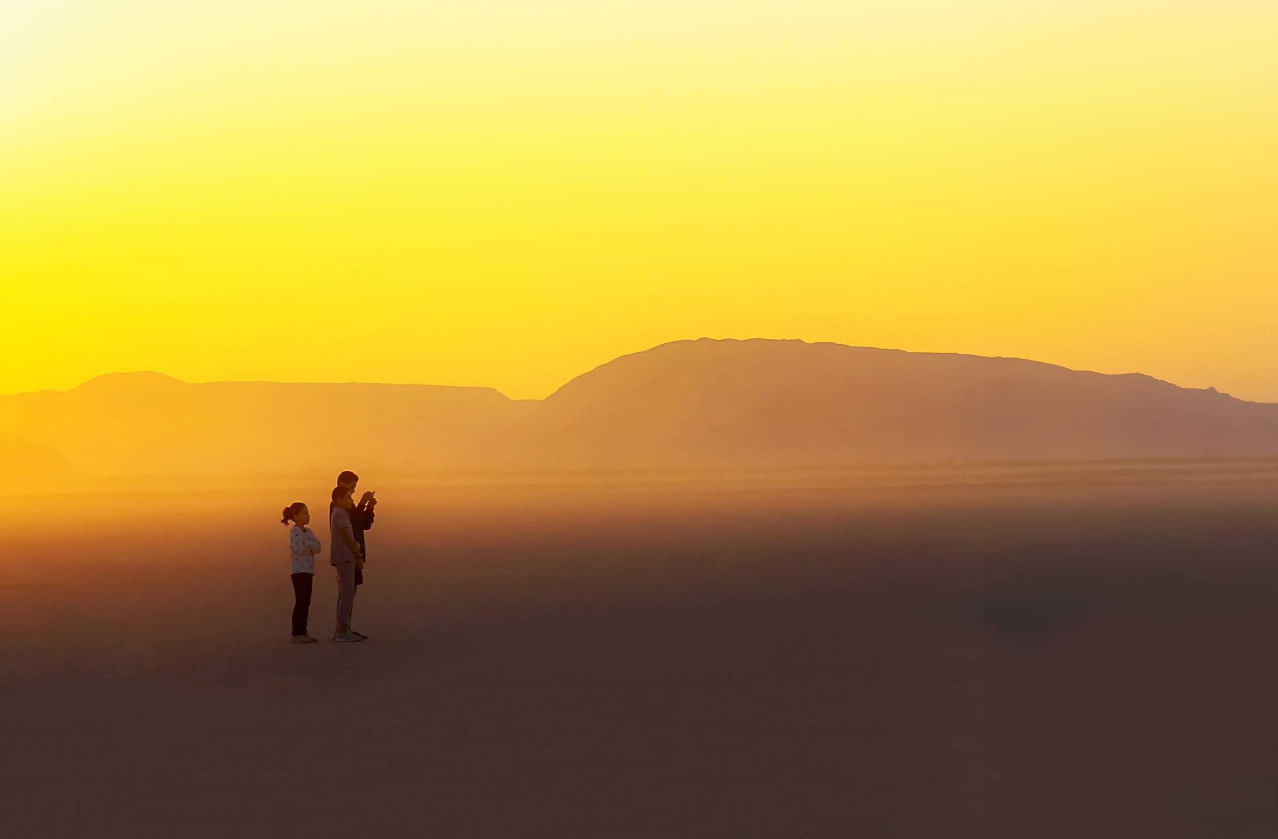 Three people standing in profile on a vast desert floor against a bright yellow sunset sky, with layered mountain silhouettes in the background.