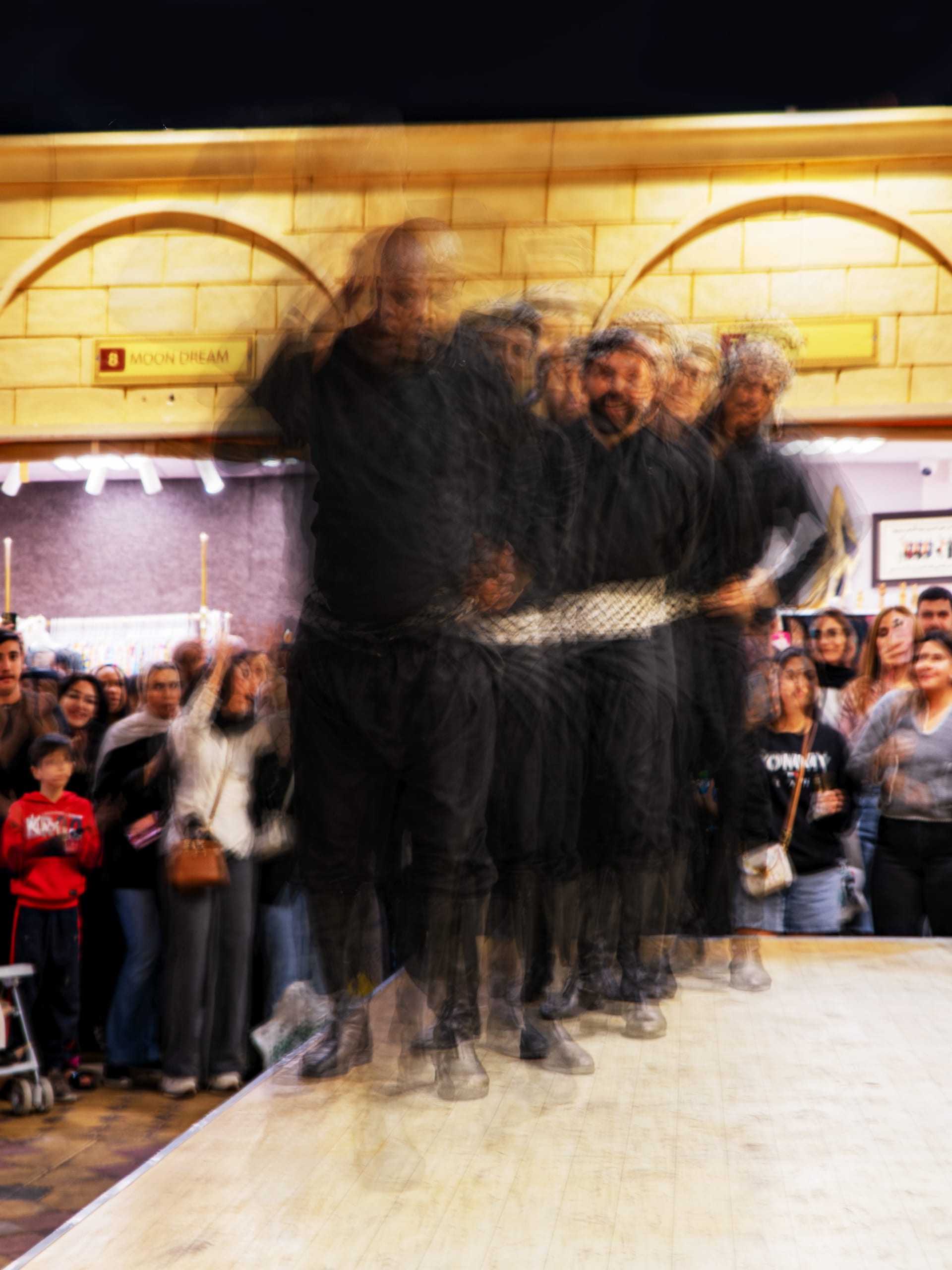 A group of male dancers in black clothing and patterned sashes perform on a stage with a slow-shutter motion blur effect, while an audience watches from the left and right sides.