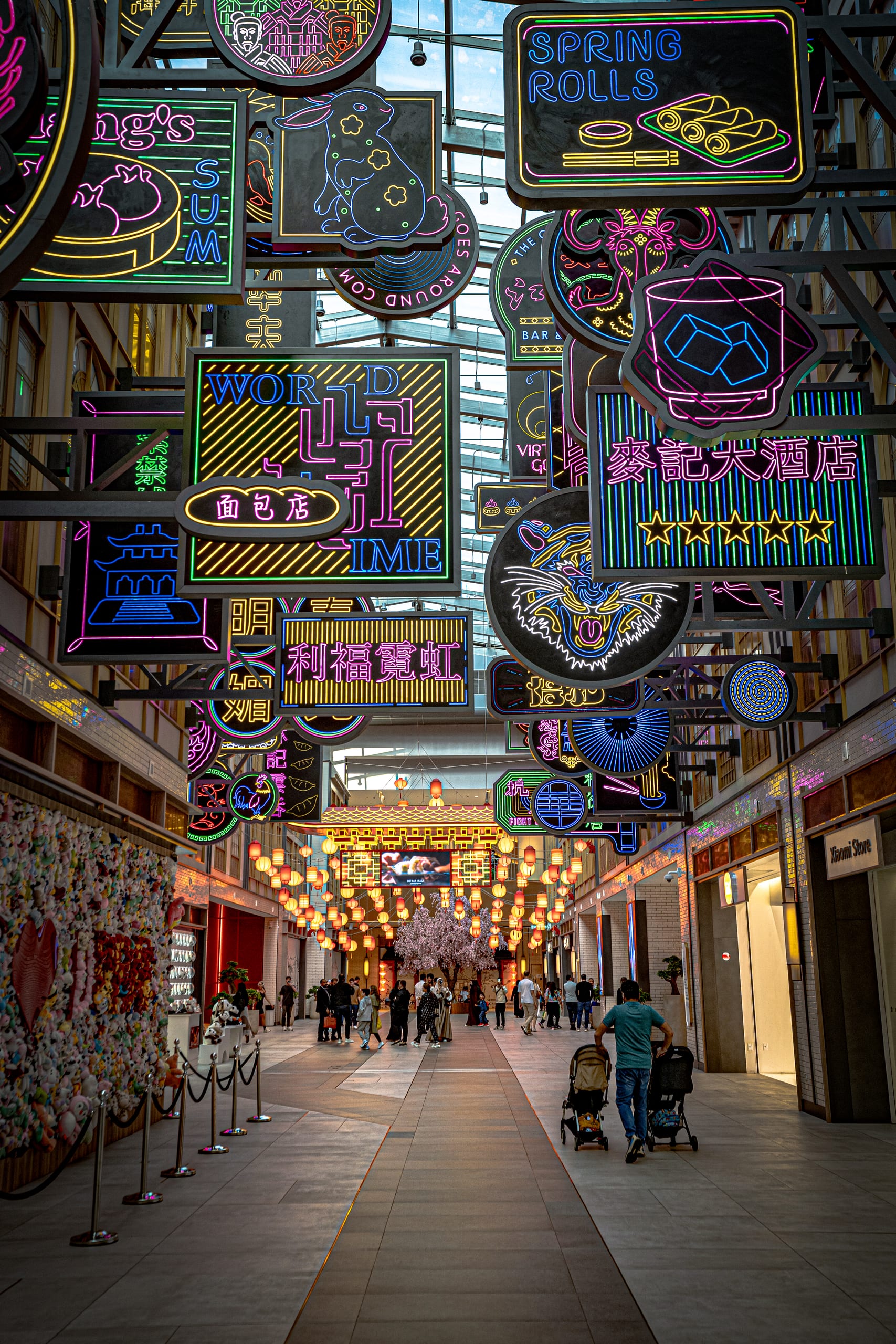 Low-angle shot looking down an indoor arcade crowded with glowing neon signs suspended overhead, with shoppers walking below and traditional lanterns lining the distance.