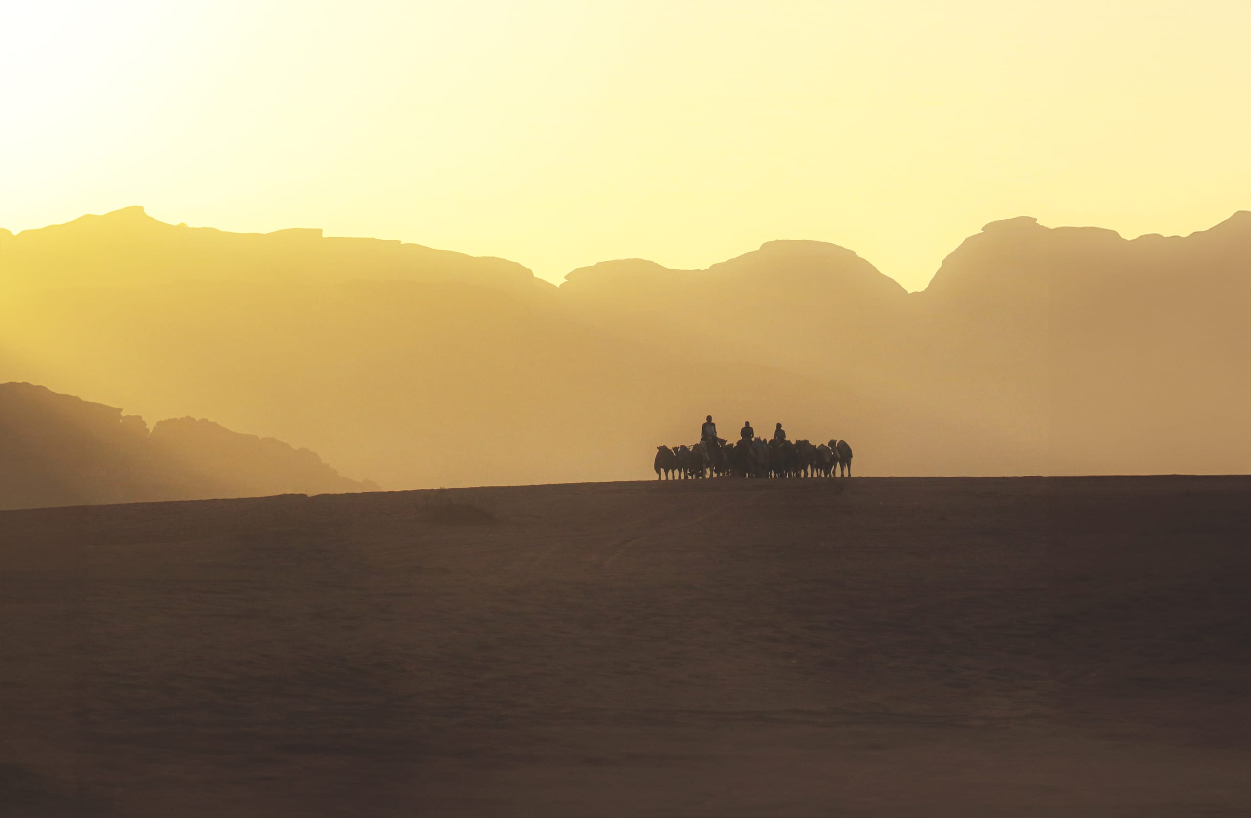 Silhouette of a caravan of people riding camels crossing a dark desert ridge in front of a hazy golden sunset and distant mountain silhouettes.